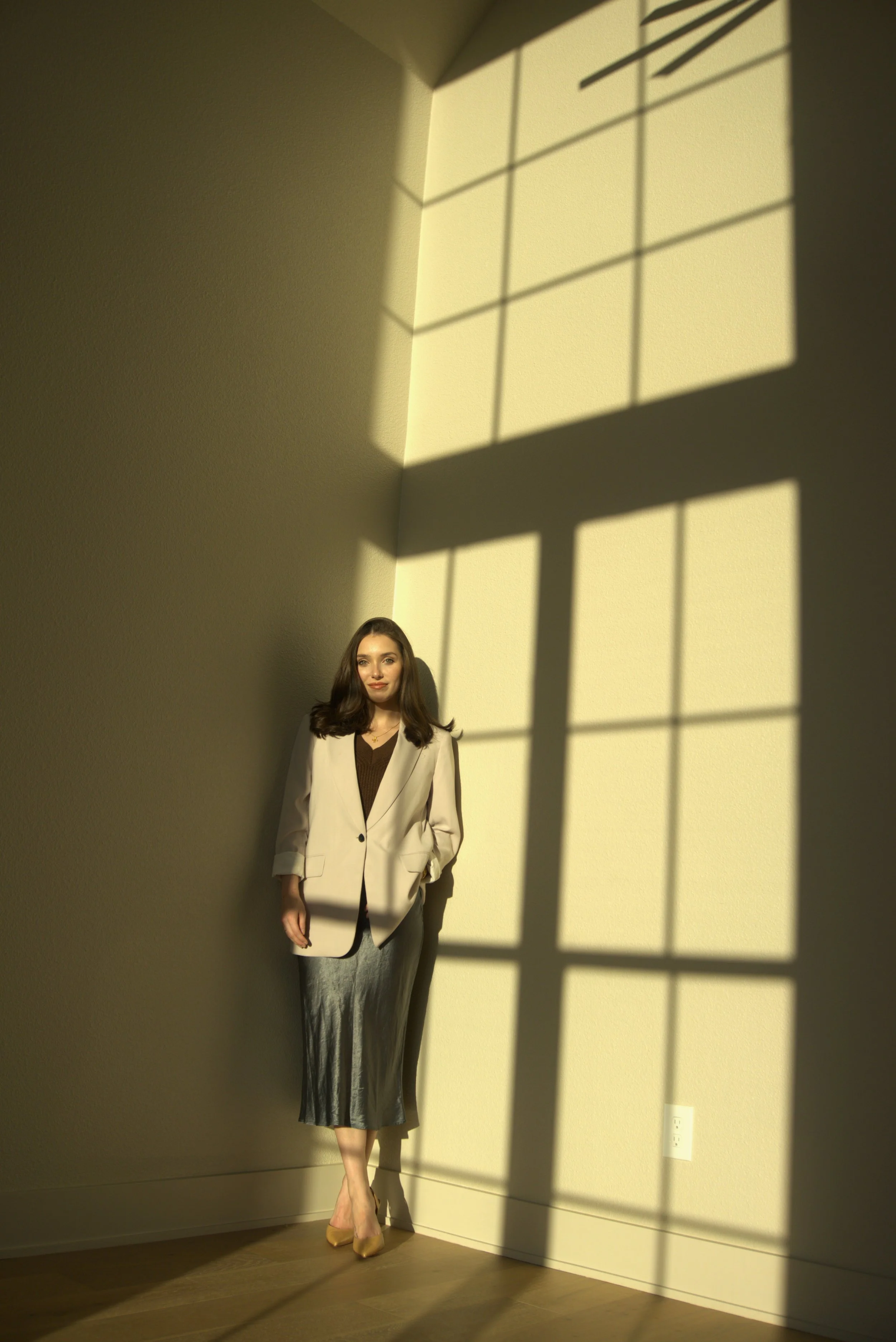 A woman in a cream blazer, black top, and silver skirt standing in a sunlit room, cast shadow of a window grid on the wall.