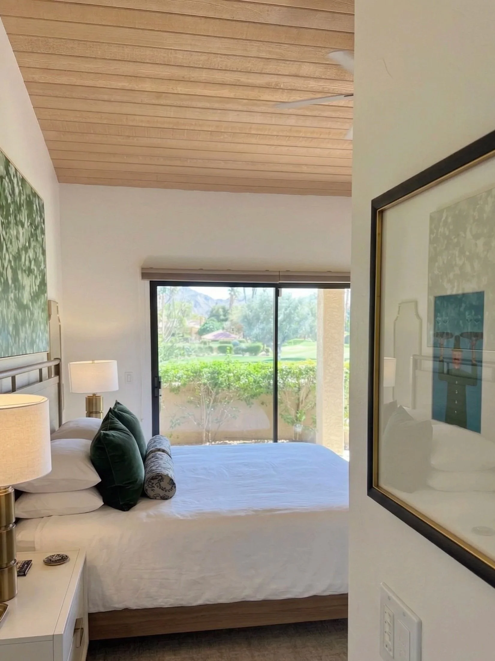 A bedroom with a bed, pillows, lamps, and a sliding glass door opening to a view of a garden or park.