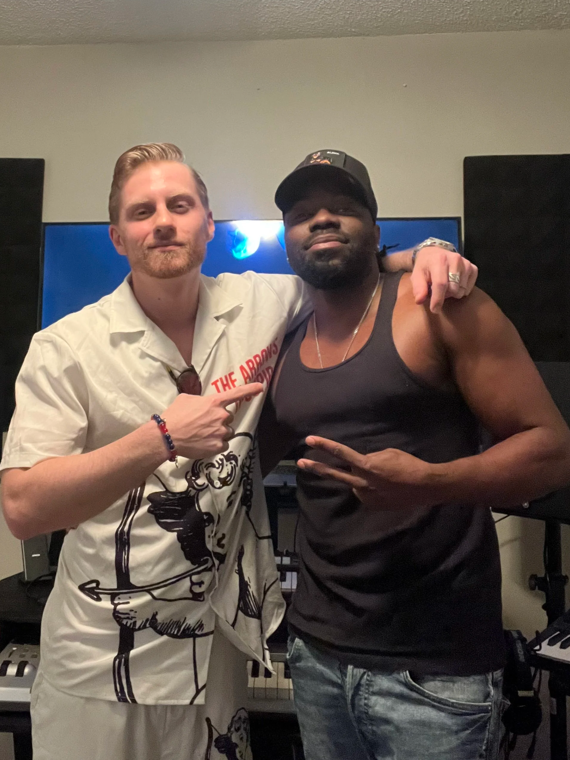 Two men posing together in a room with musical equipment. One man is wearing a white shirt, and the other man is wearing a black tank top and a baseball cap. They are standing close, with the man in the white shirt pointing at the other man.