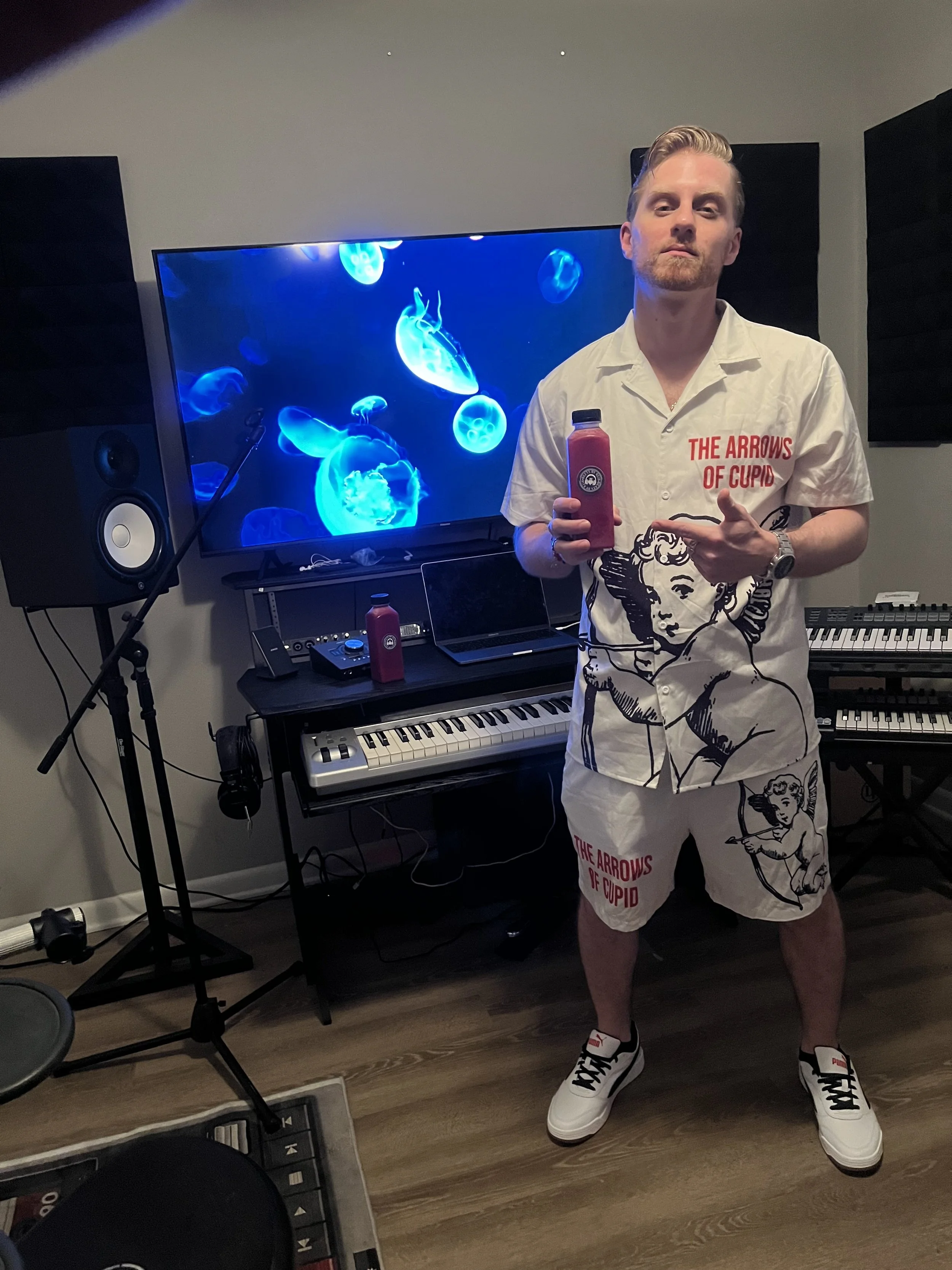 A young man with light hair and a beard standing in front of music equipment, holding a red bottle, wearing a white shirt and shorts with 'The Arrows of Cupid' printed on them, and white sneakers with black and red accents.