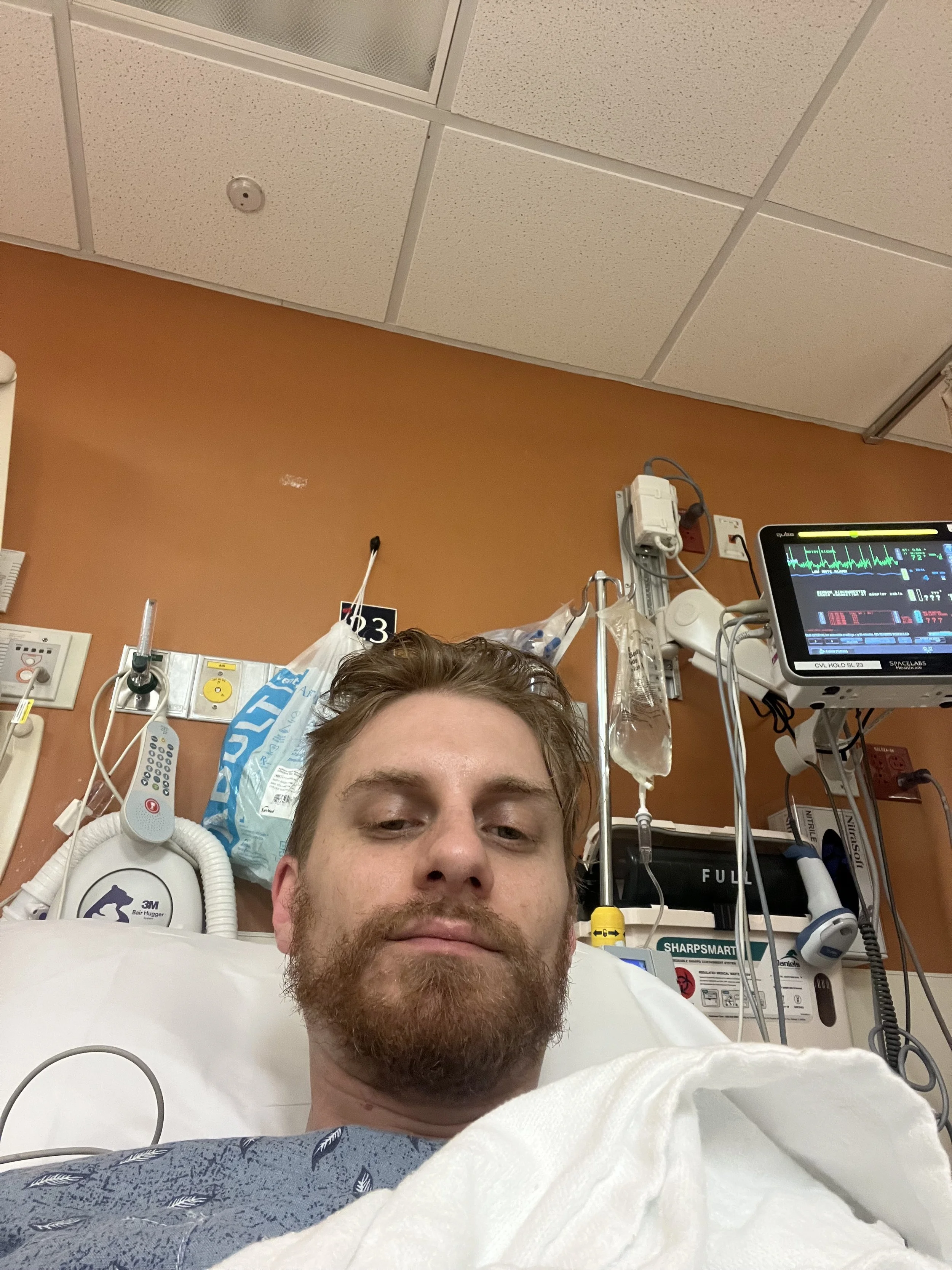 A man with a beard and mustache lying in a hospital bed, taking a selfie. Medical equipment and monitors are visible around him, and he appears to be in a hospital room.