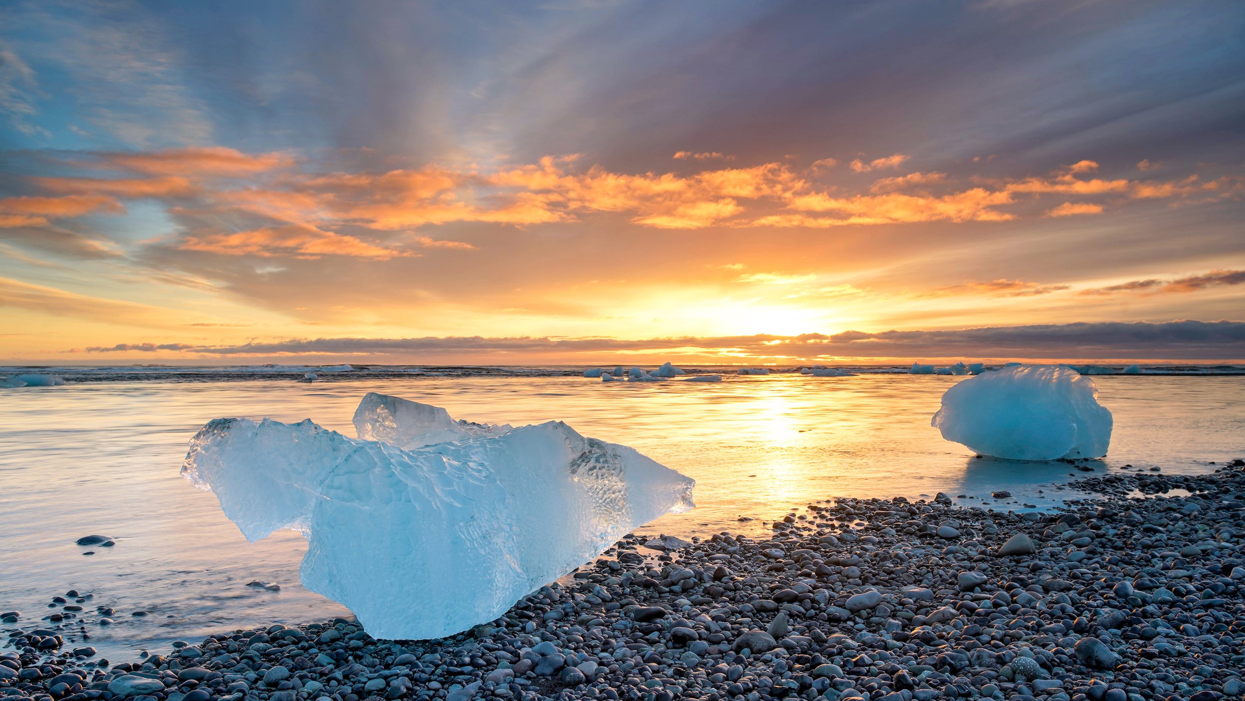 Icebergs on a rocky beach during sunset with colorful sky and calm water.