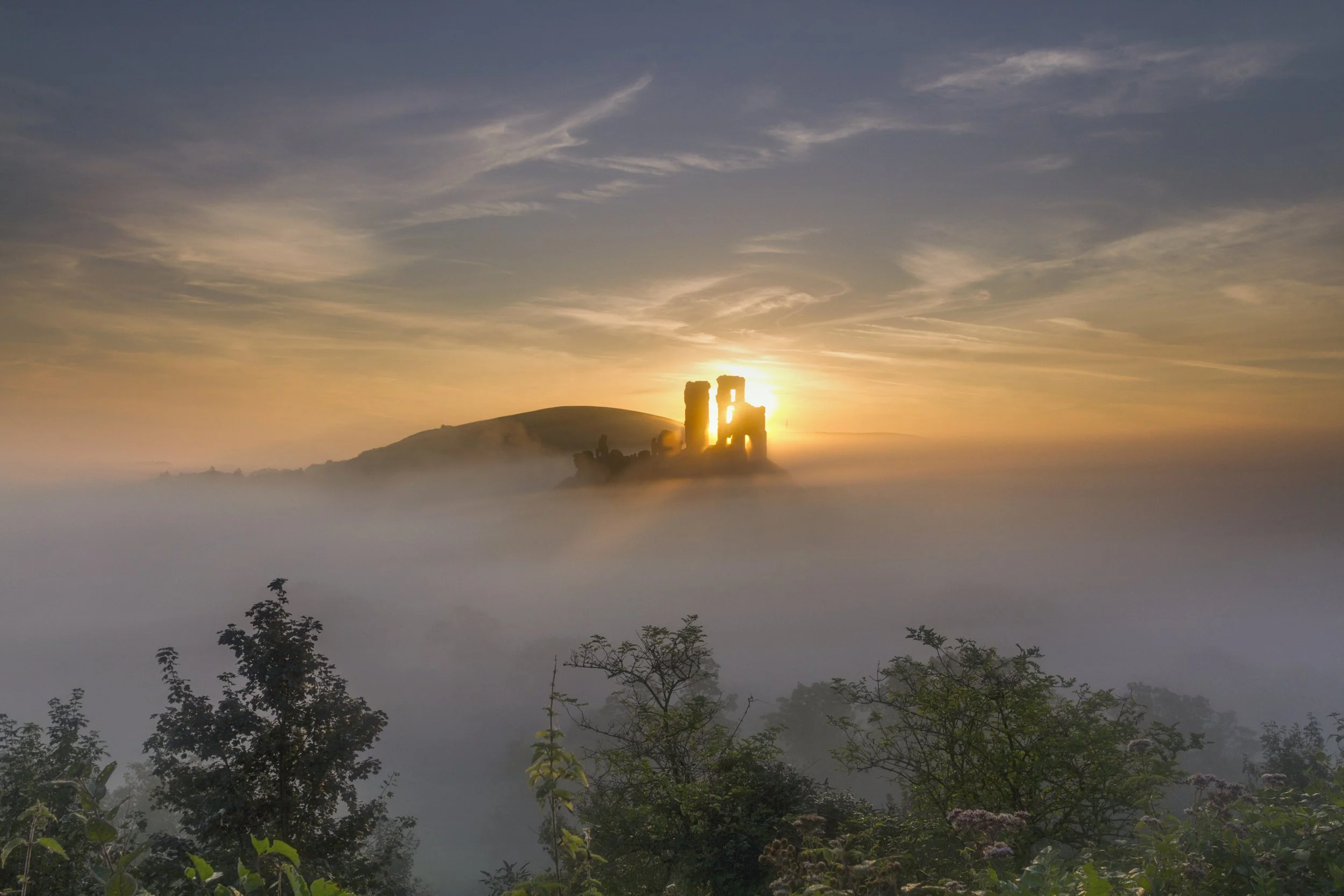 Sunset over a foggy landscape with ancient ruins on a hill and trees in the foreground.