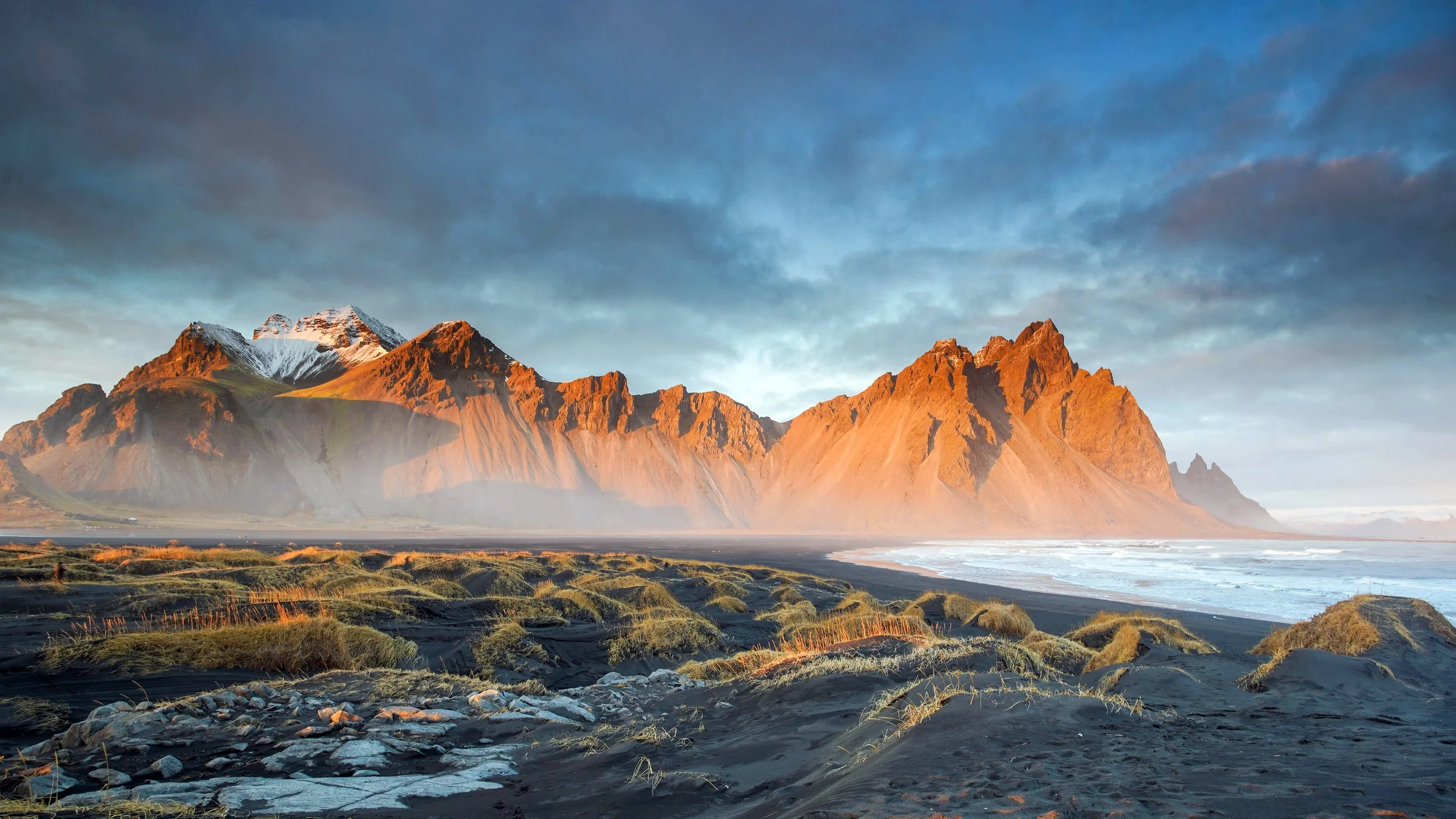 Scenic view of a rugged coastline with black sand, grass-covered dunes, and dramatic orange-tinted mountains under a cloudy sky.