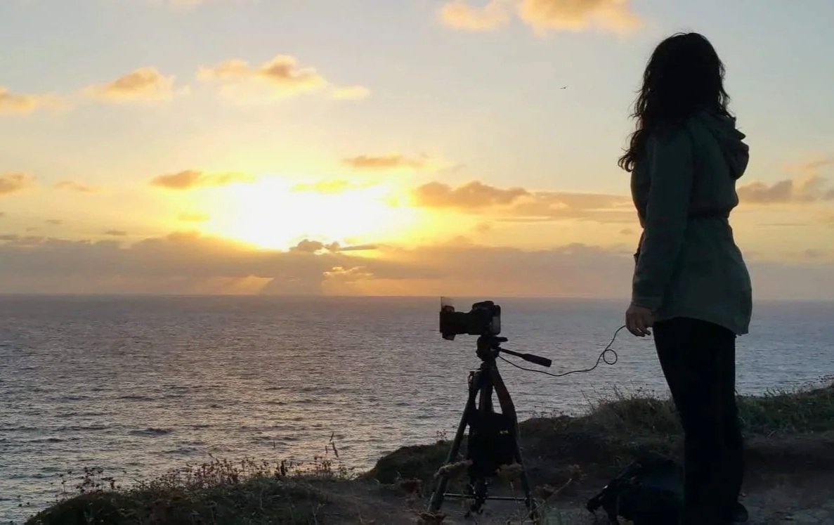 A woman standing near a camera on a tripod, overlooking the ocean during a sunset, with clouds and a setting sun in the sky.
