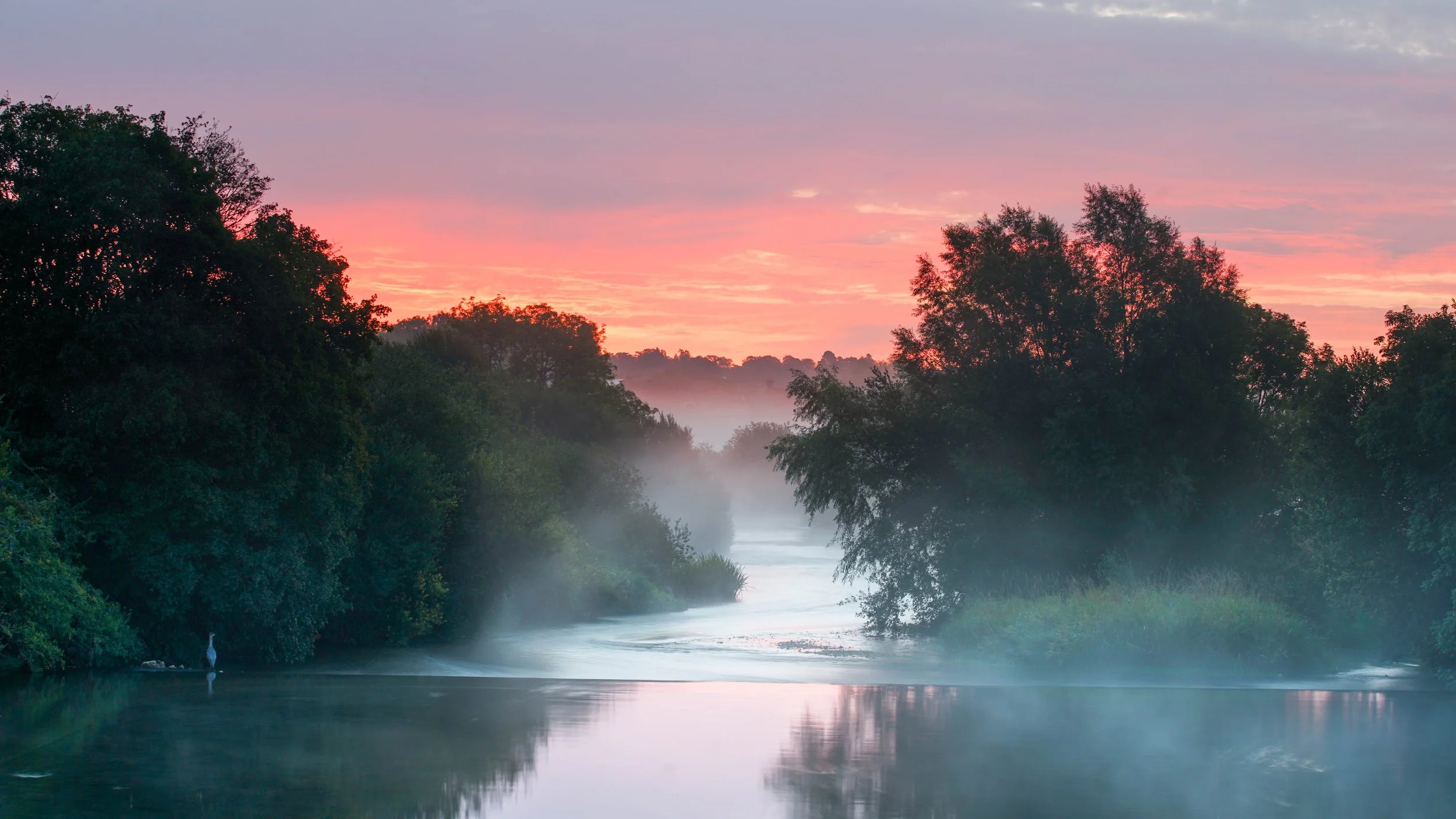 A serene river scene at dawn with mist rising over calm water, surrounded by lush trees and a colorful pink and orange sky.