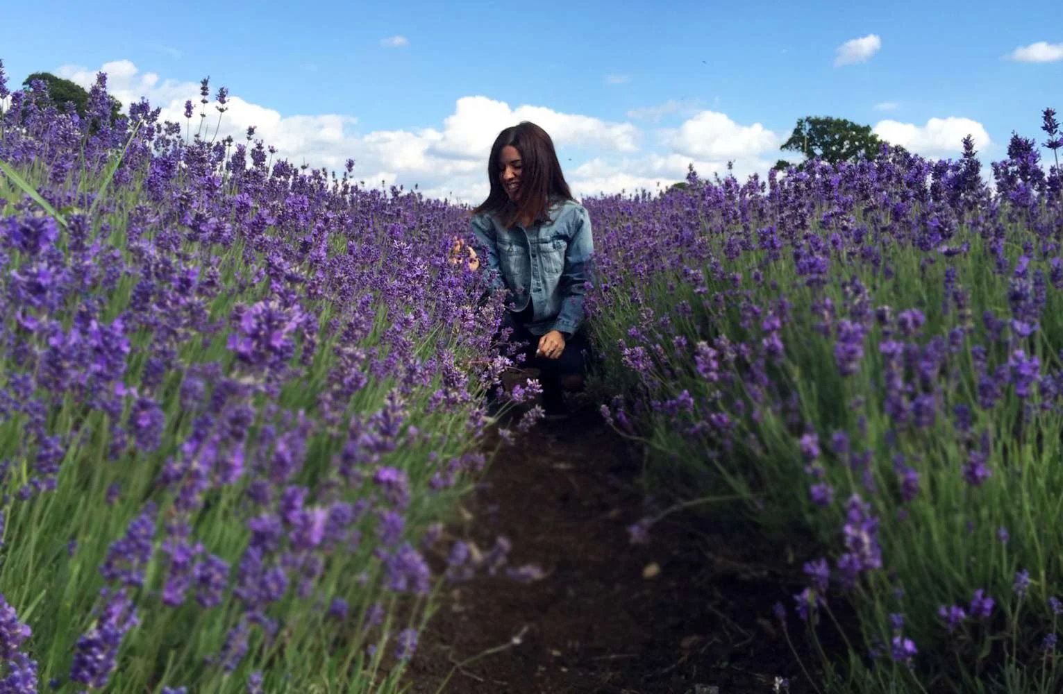 A woman in a denim jacket is kneeling in a field of purple lavender flowers under a blue sky with scattered clouds.