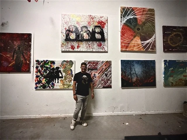 A man stands in front of a white gallery wall displaying nine abstract paintings with vibrant colors and dynamic brushstrokes.