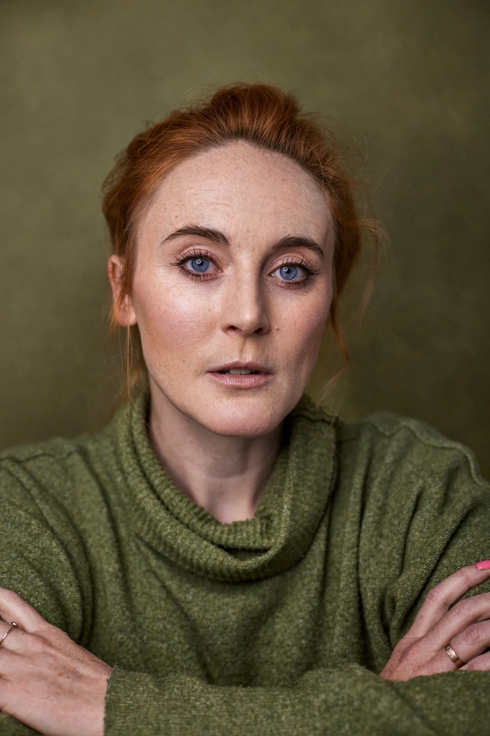 A woman with red hair and blue eyes wearing a green sweater, looking at the camera, with a neutral background.