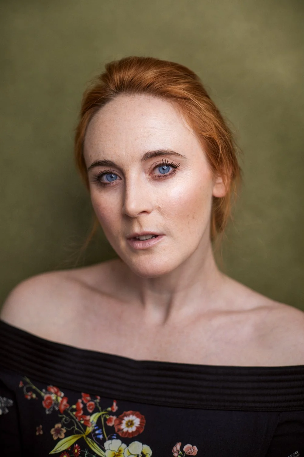 A woman with red hair and blue eyes, wearing an off-the-shoulder black top with floral embroidery, looking directly at the camera against a muted green background.