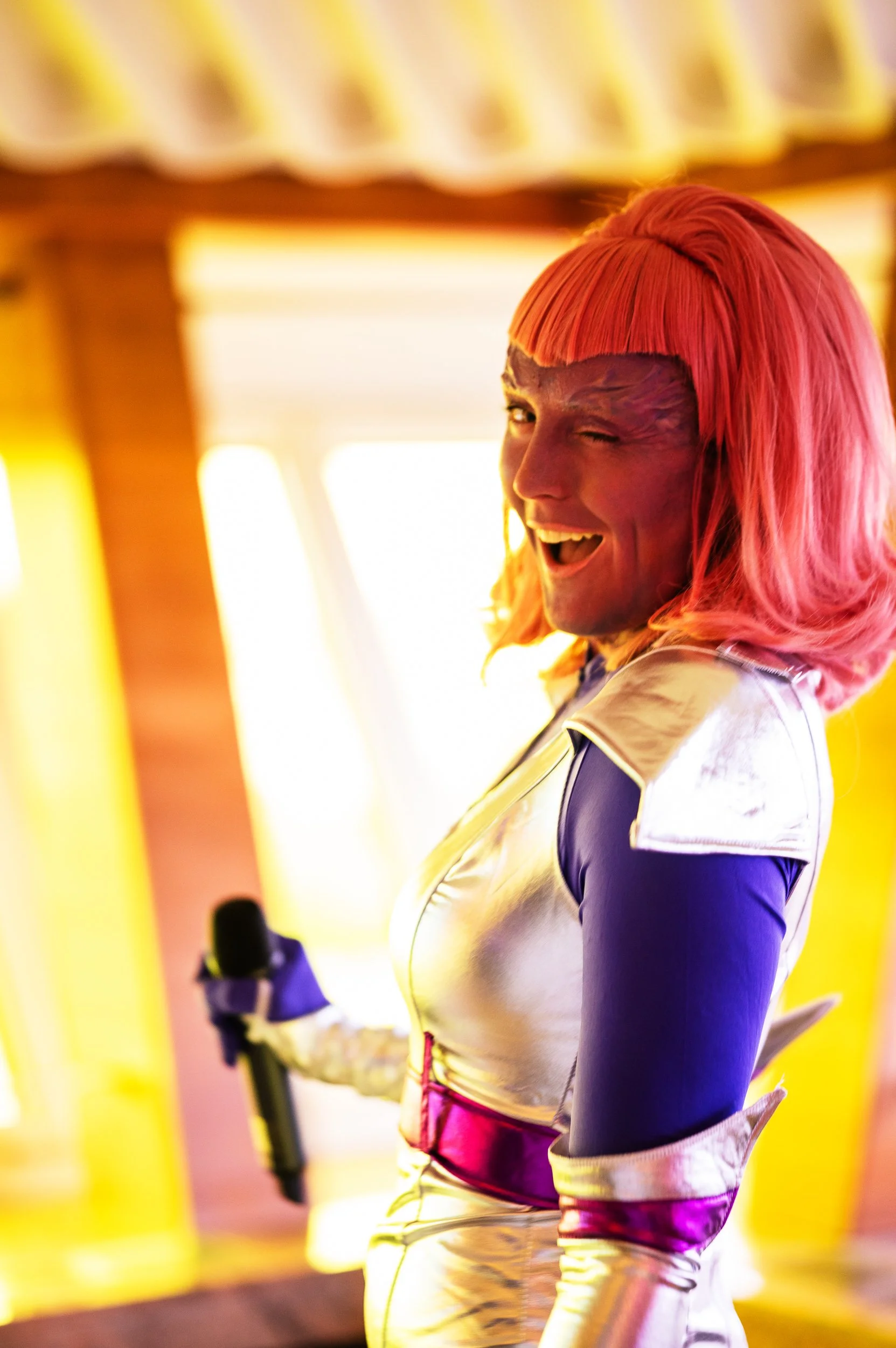 Person dressed as a superhero with a vibrant red wig, winking and smiling while holding a microphone, indoors with yellow tones and large windows in the background.
