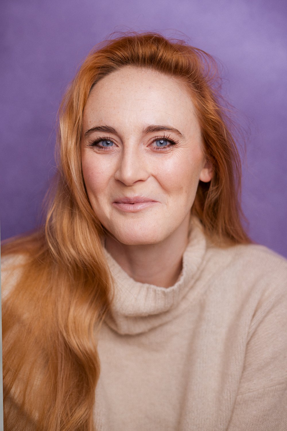 A woman with long red hair, blue eyes, and fair skin, wearing a beige turtleneck sweater, smiling slightly against a purple background.