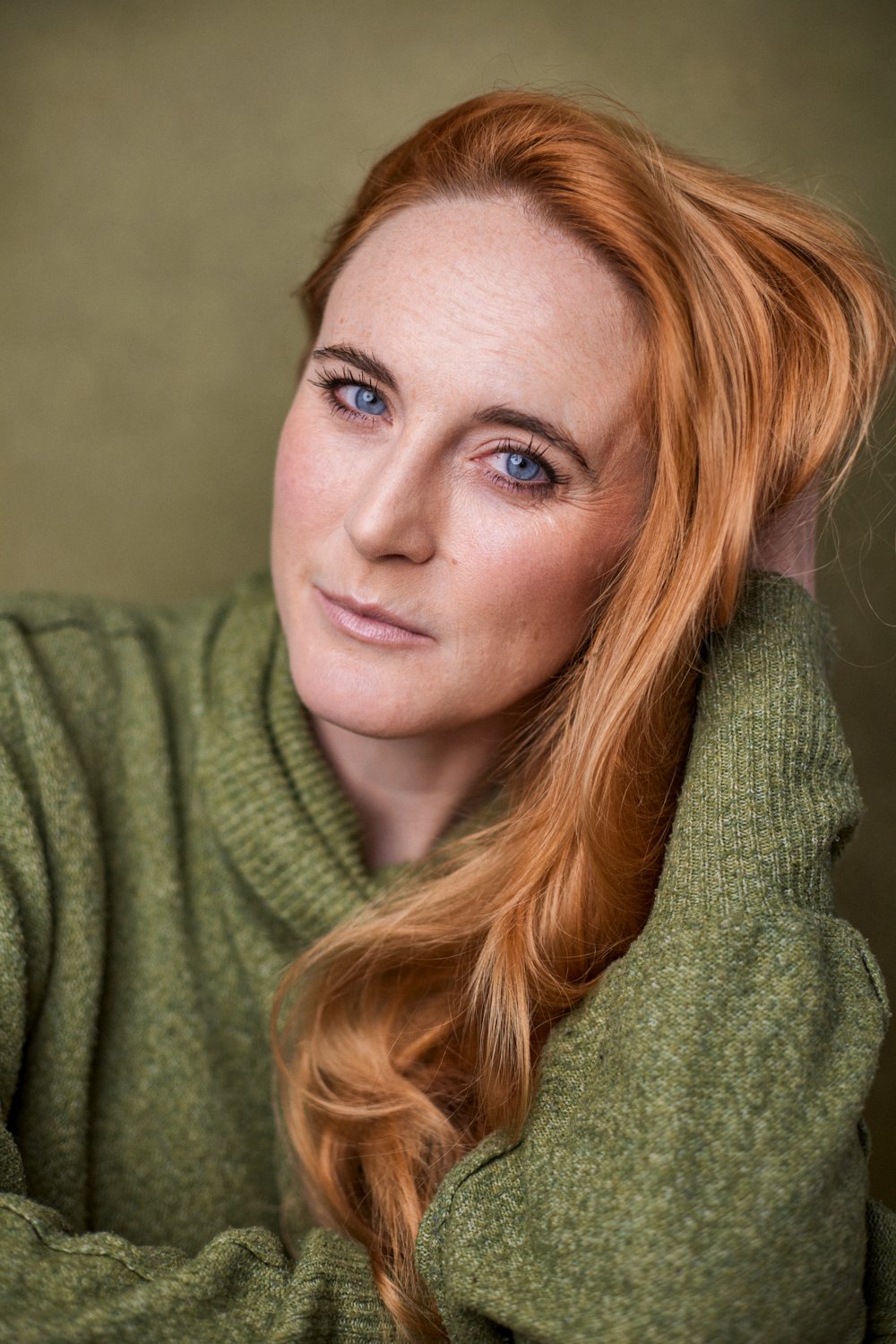 A woman with long red hair and blue eyes wearing a green sweater, looking at the camera with a gentle expression against a neutral background.