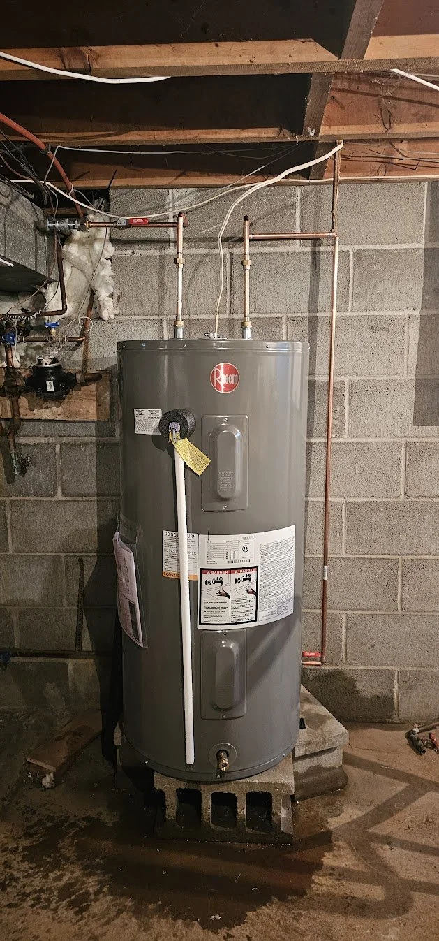 A Rheem water heater installed on concrete blocks in a unfinished basement with concrete walls and wood ceiling. Piping and wiring are connected to the water heater.