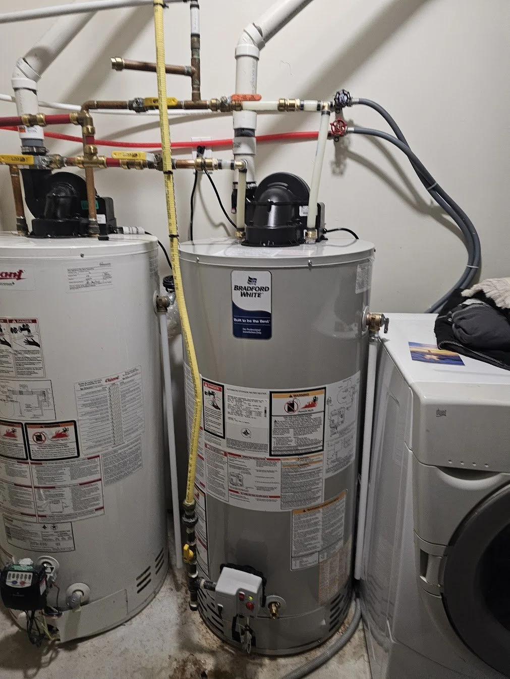 Indoor plumbing utility room featuring a water heater on the left, another water heater in the middle, and a washing machine on the right, with various pipes and hoses connected to the appliances.