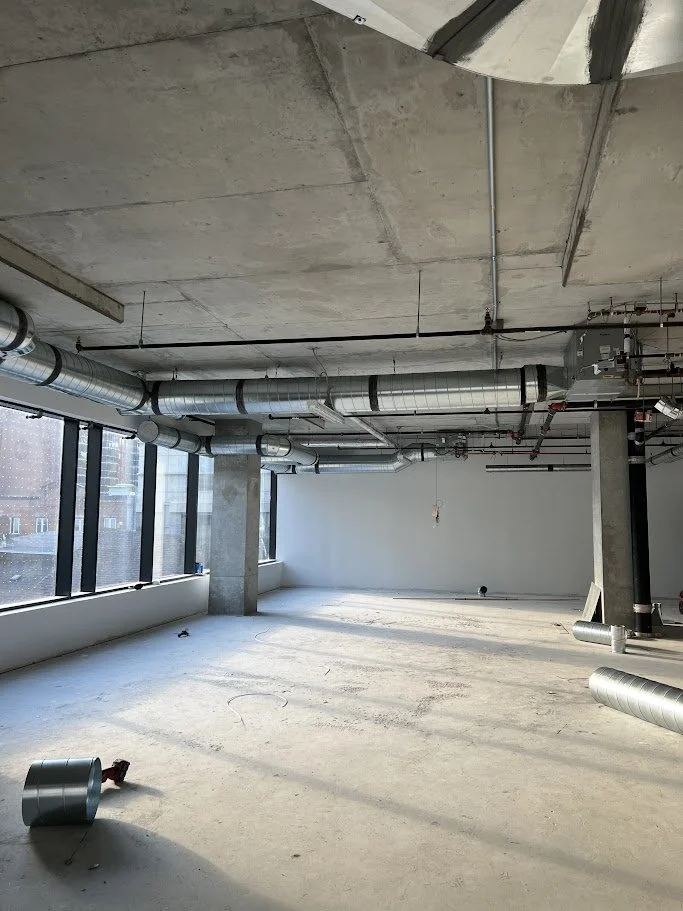 Empty unfinished commercial space with large windows, exposed ductwork and pipes, concrete floors, and no interior walls yet.