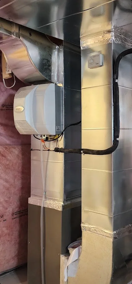 Close-up of a basement mechanical room system with metallic ducts, a white humidifier or air purifier, and electrical wiring.