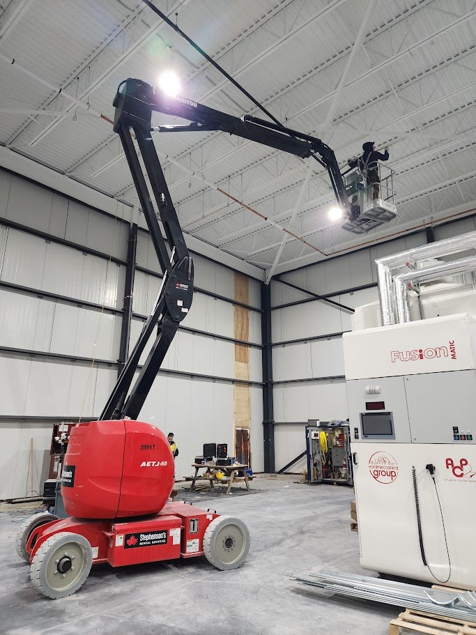A worker on a cherry picker elevated high in an industrial warehouse next to the ceiling, working on a light. The warehouse has a high ceiling with white roof panels and metal beams. There is machinery labeled Fisher Fusion on the right side and vari