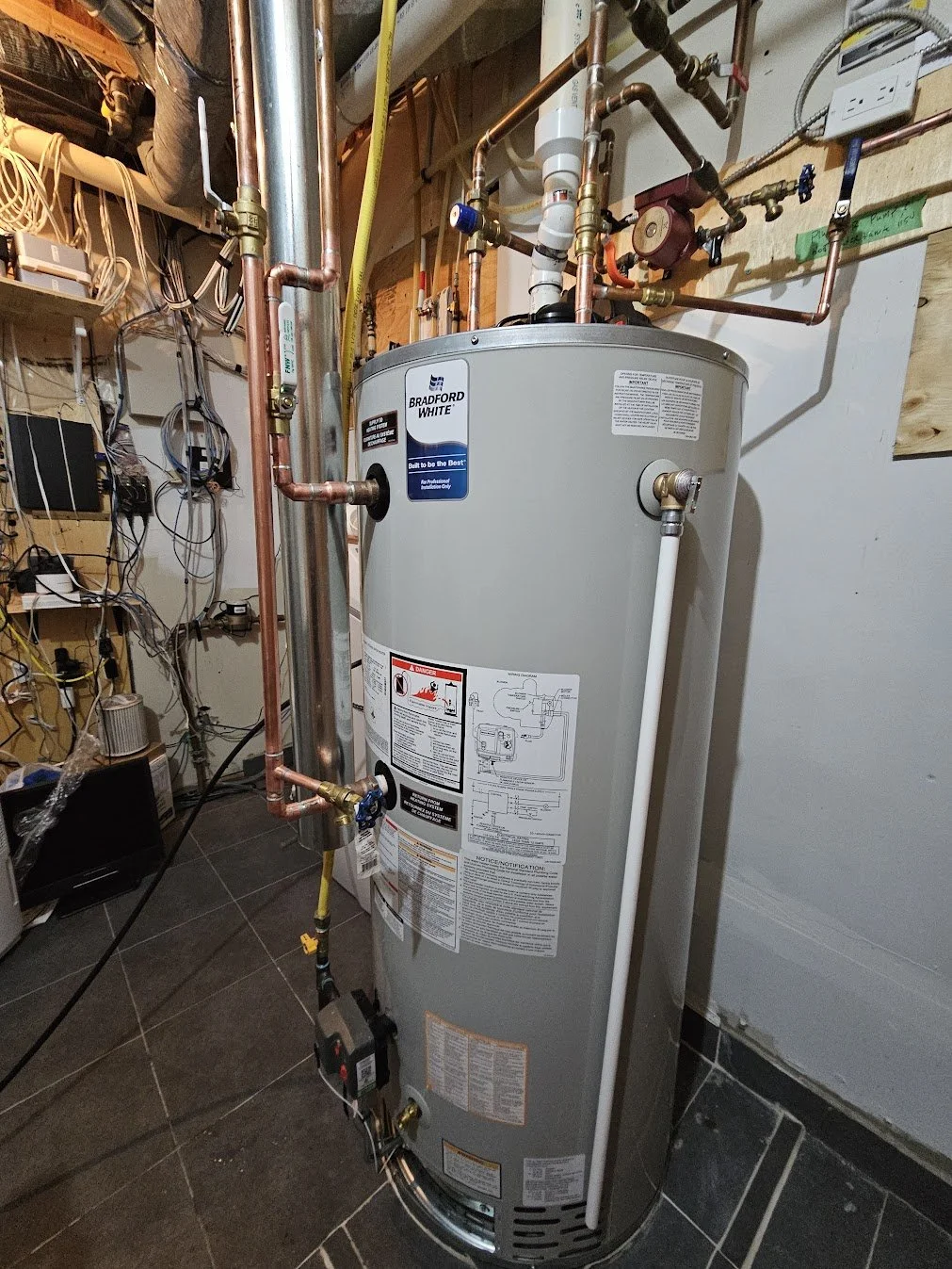 A tall, gray Bradford White water heater with various pipes, valves, and wiring connected, situated in a utility room with a tiled floor and unfinished walls with electrical wiring and shelving.