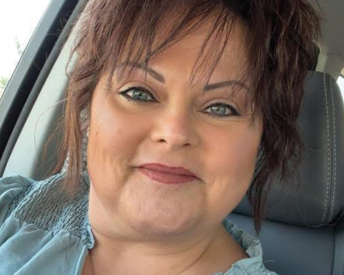 Close-up of a woman with short reddish-brown hair and blue eyes, sitting in a car, smiling.