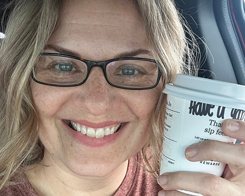 Woman with glasses smiling and holding a coffee cup inside a car.
