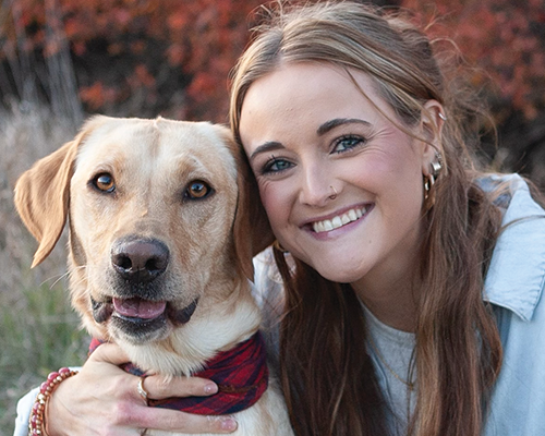 A young woman with long brown hair smiling and hugging a yellow Labrador Retriever outdoors.