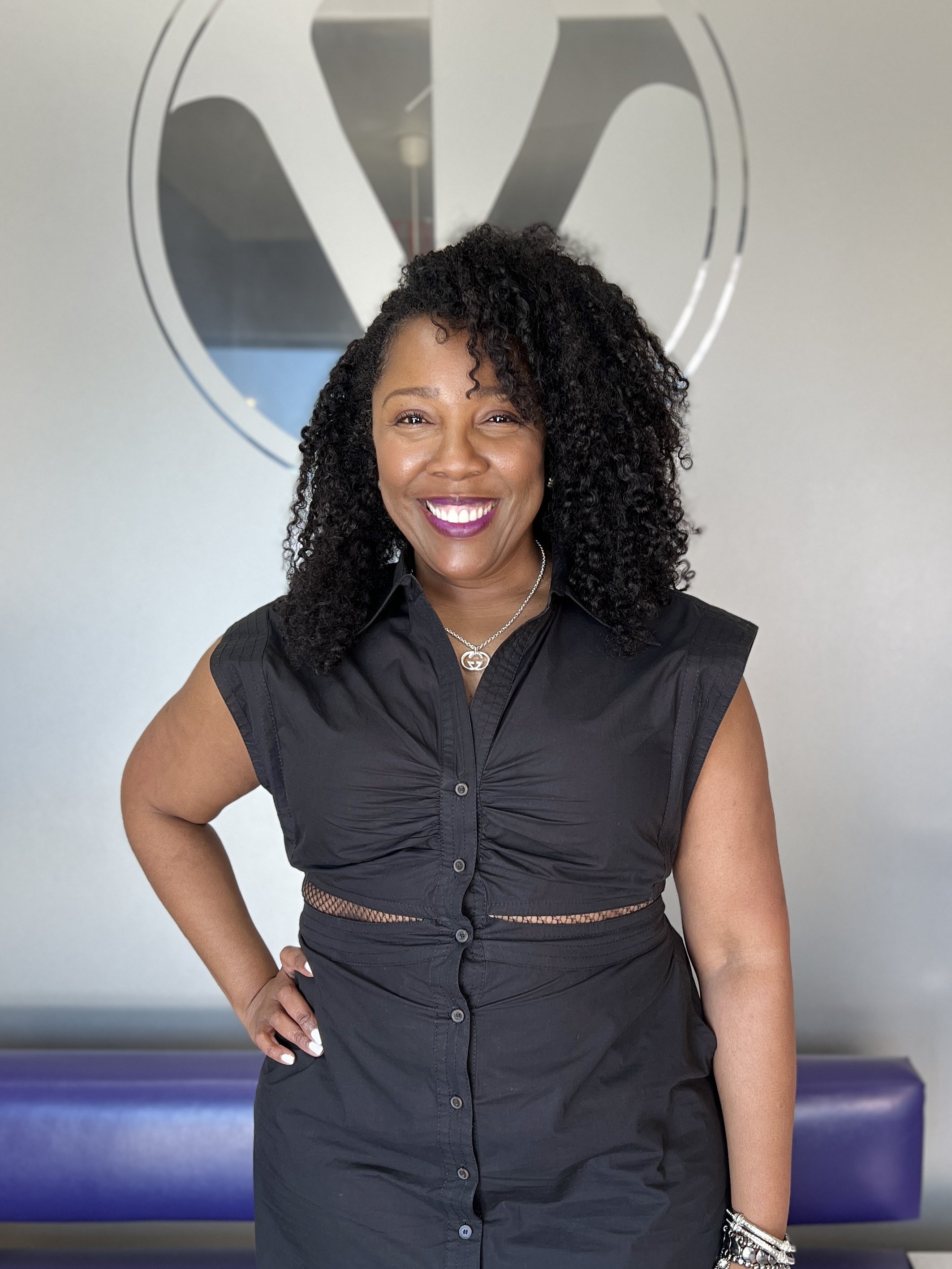 A woman with curly black hair and a bright smile, wearing a sleeveless black dress with buttons down the front, standing in front of a wall with a logo in the background.