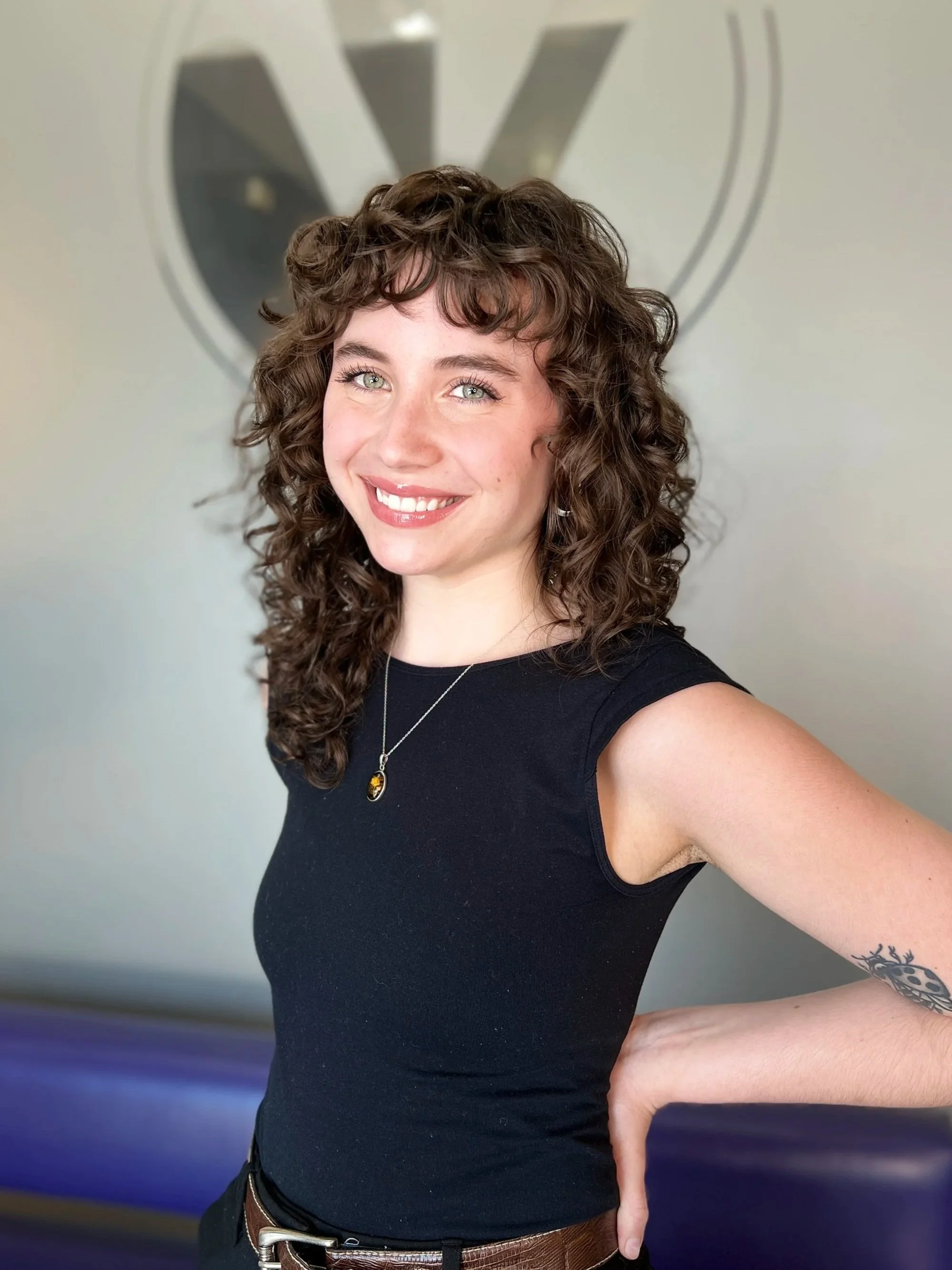 A woman with curly brown hair, light skin, and blue eyes is smiling and looking at the camera. She is wearing a black sleeveless top, a necklace with a pendant, and has a tattoo on her right arm. She is standing in front of a blurred background with a logo or emblem.