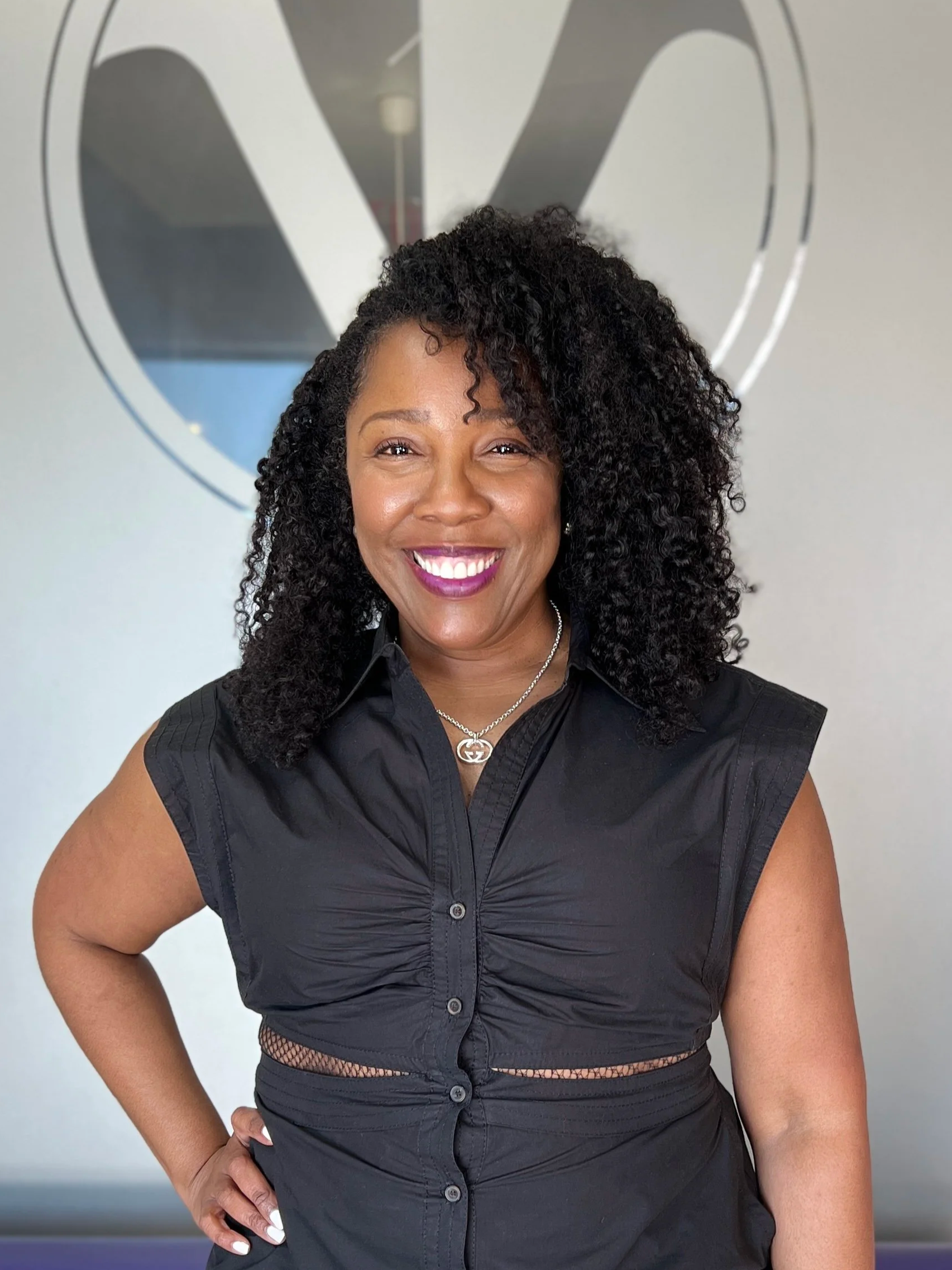 A woman with curly black hair, wearing a black sleeveless shirt and a silver necklace, smiling and posing in front of a wall with a large logo.