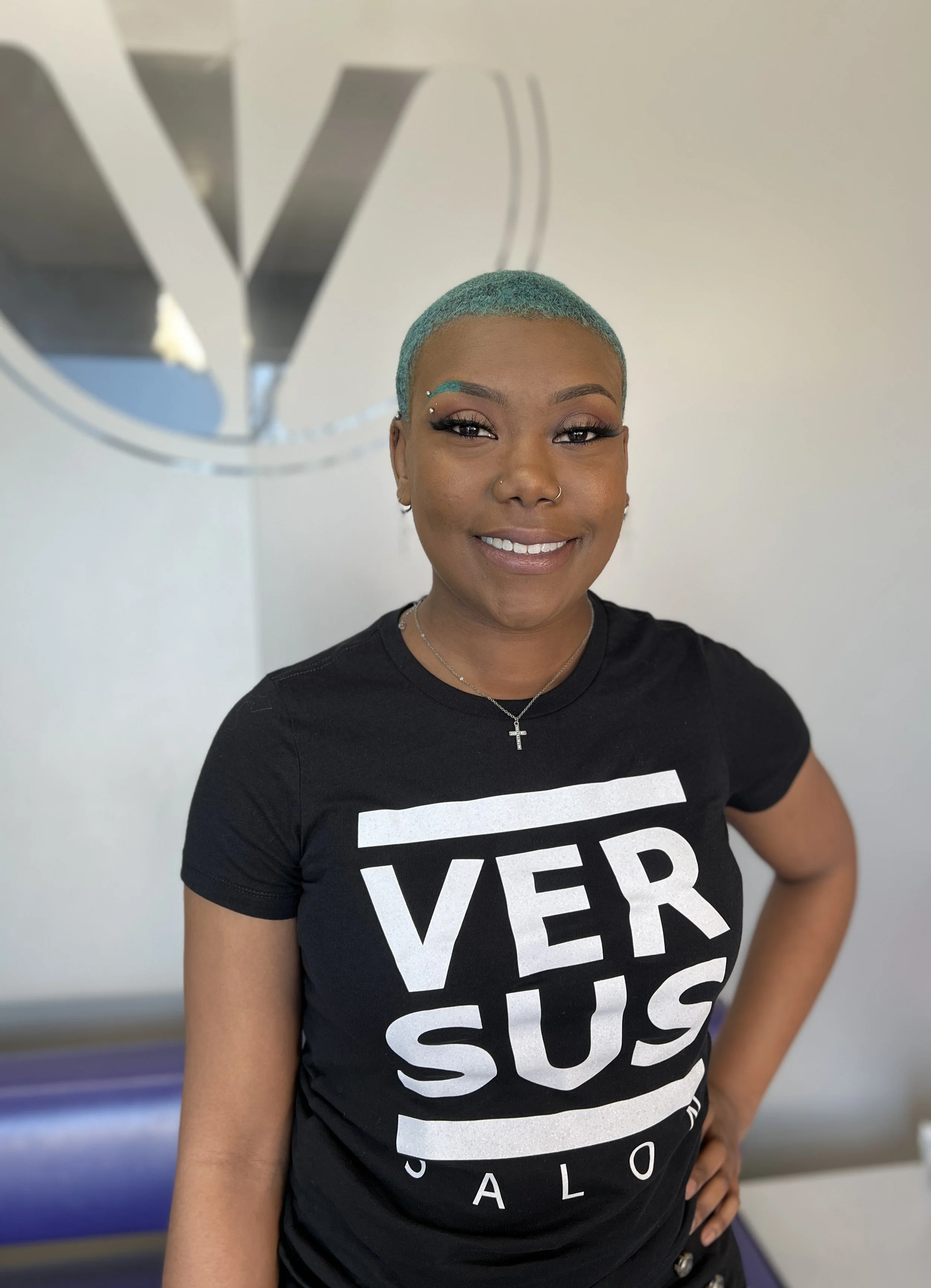 A woman with short turquoise hair smiling, standing indoors in front of a wall with a logo and wearing a black t-shirt with white text.