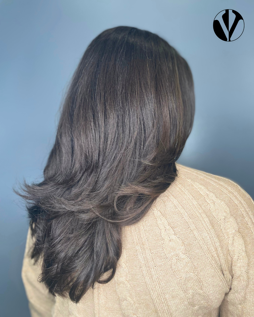 Back view of a woman with dark brown, wavy, shoulder-length hair wearing a beige sweater against a gray background.