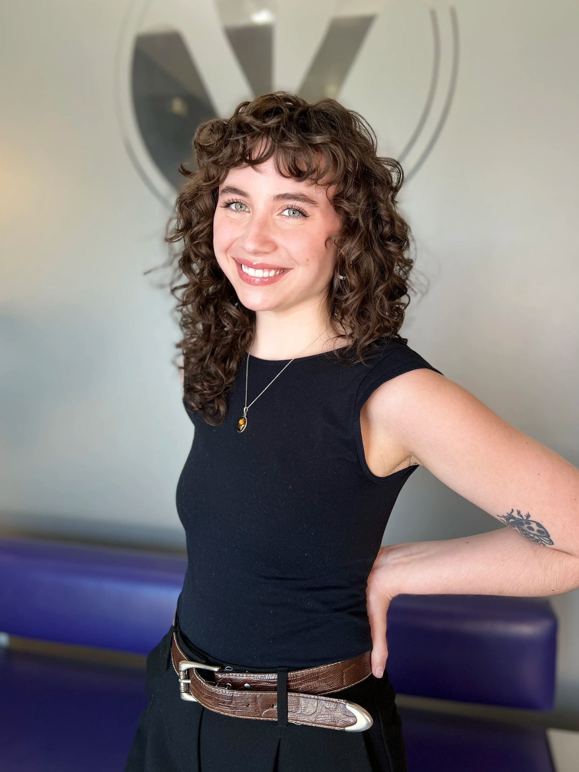 A young woman with curly brown hair, blue eyes, and light skin, smiling and wearing a black sleeveless top, a silver necklace, and a brown belt, standing in front of a wall with a large logo.