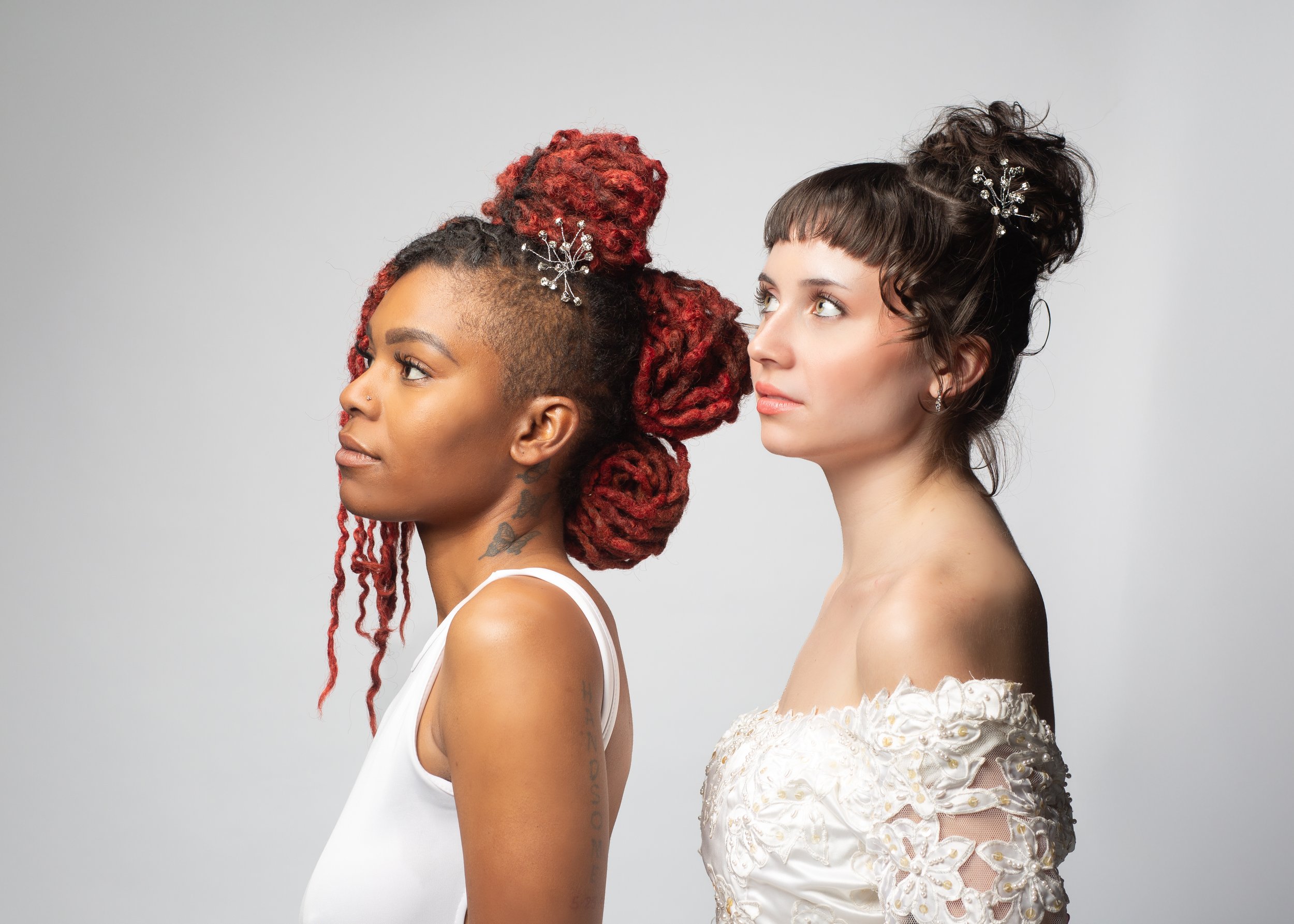 Two women with styled hair, one with red dreadlocks and the other with dark, curly hair, looking to the side on a plain background.