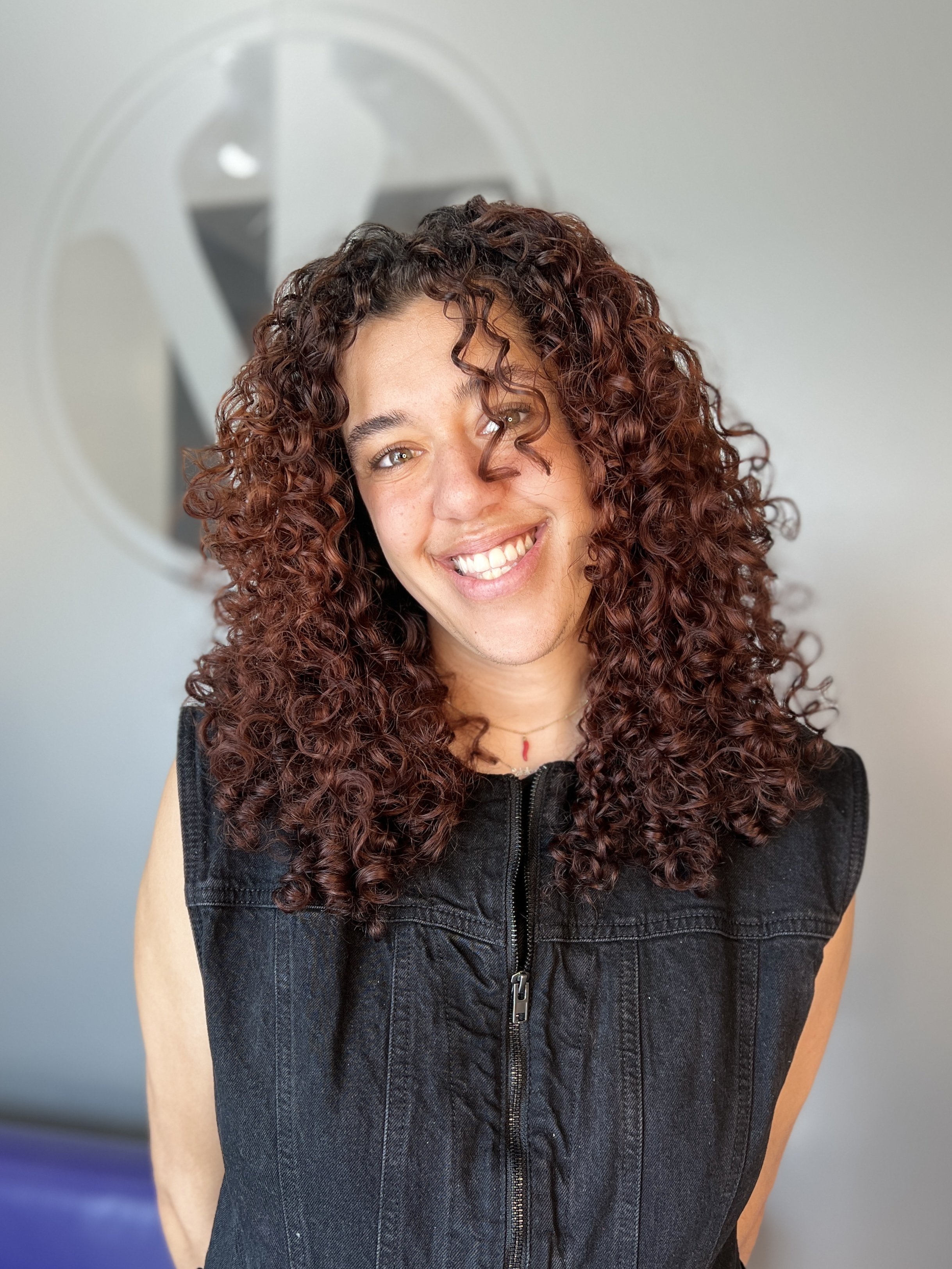 A young woman with curly red hair smiling in front of a plain wall and a circular mirror.