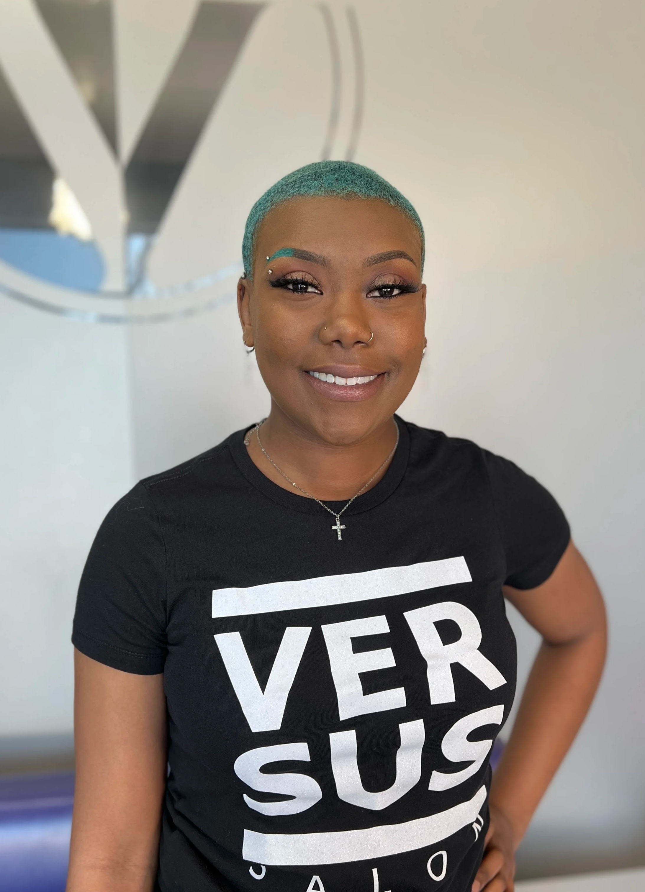 A smiling woman with short, teal hair, wearing a black T-shirt with partially visible white text, a small cross necklace, and earrings, standing in front of a white wall with a blurred logo.