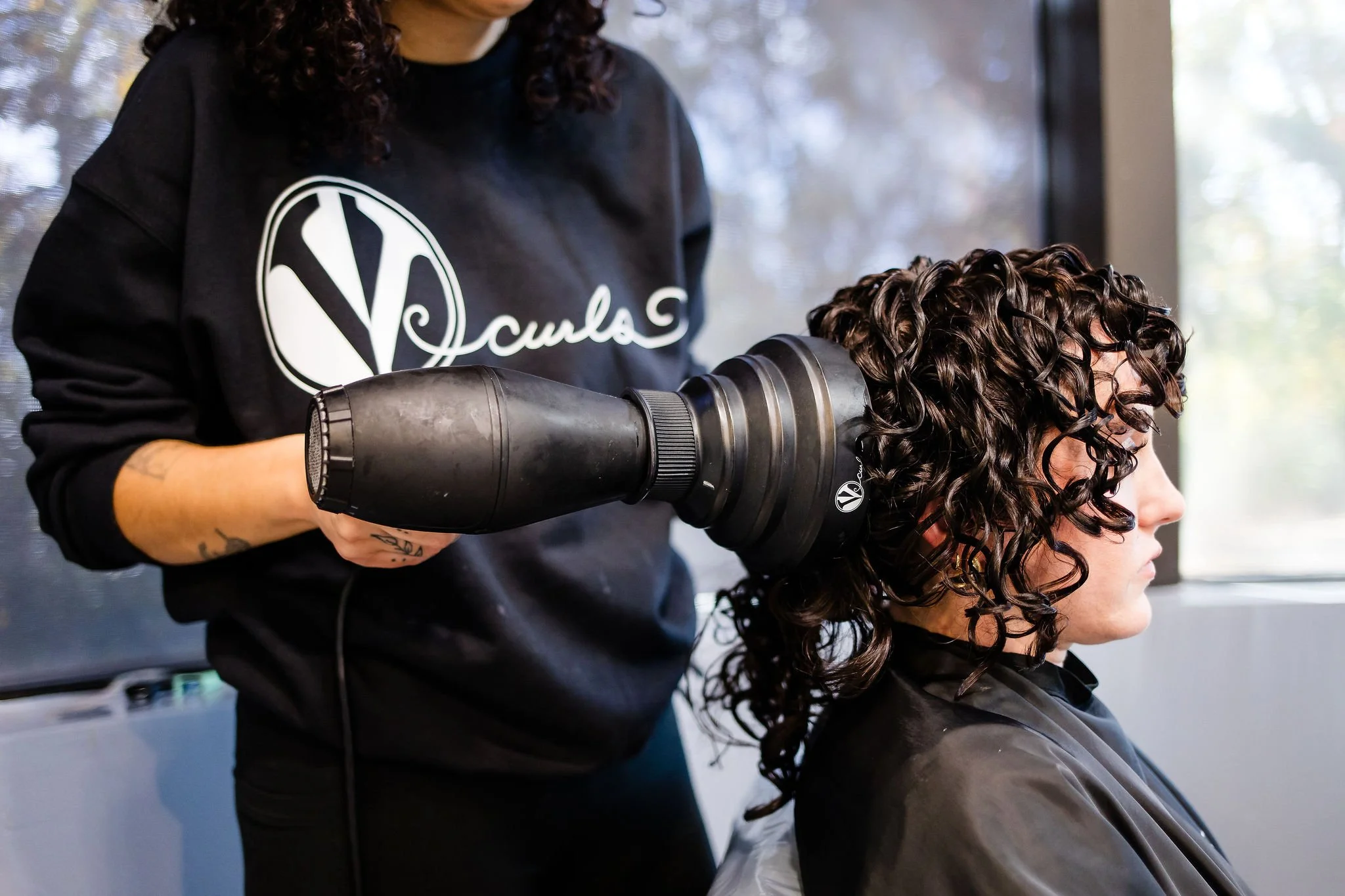 A woman with curly dark hair receiving a hair drying treatment at a salon with a specialized blow dryer.