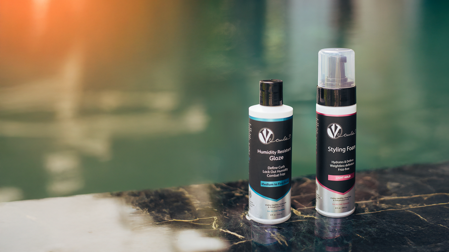 Two bottles of hair styling products, one labeled 'Humidity Resistant Glaze' and the other 'Styling Foam,' placed on a dark marble surface with a blurred water background.