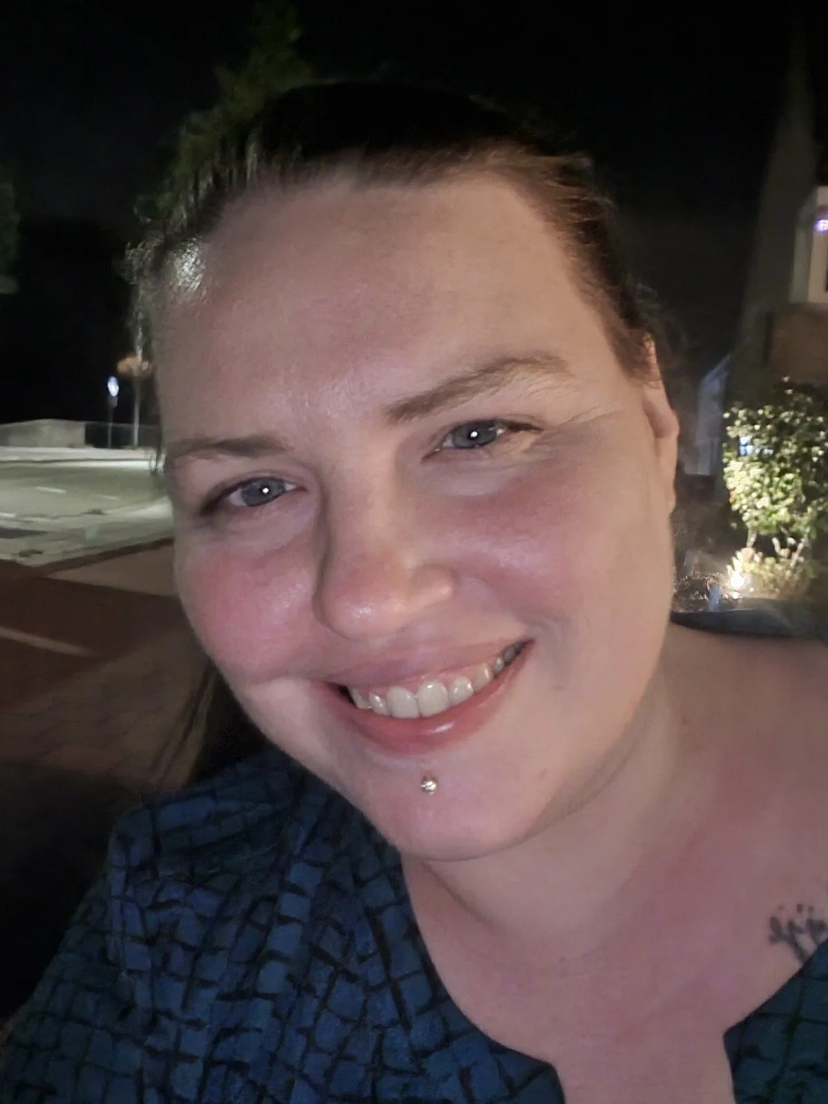Vrouw glimlacht naar de camera; avondomgeving met verlichting en groen op de achtergrond.