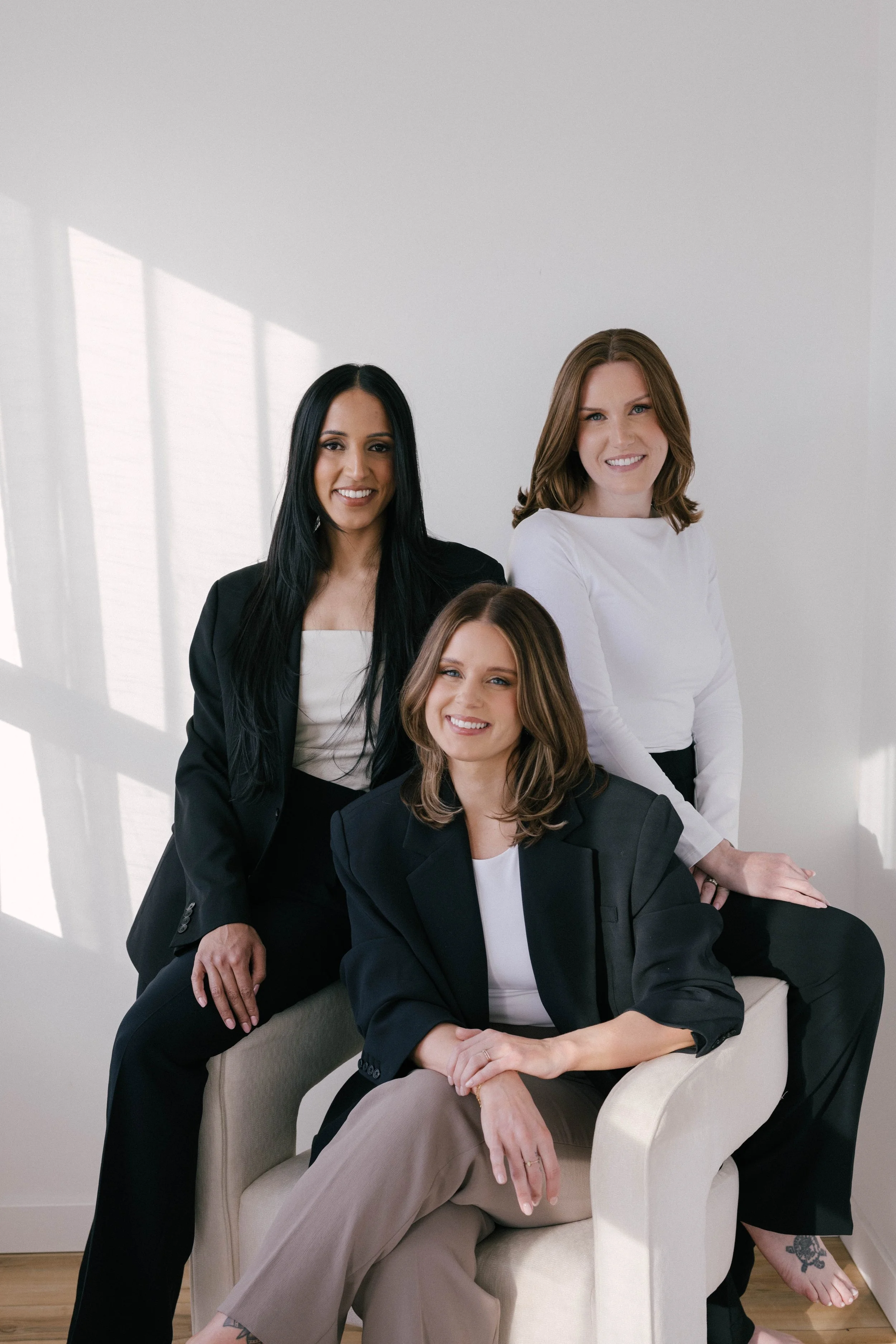 Three women smiling at the camera, one sitting on a white chair, all dressed in black and white business attire
