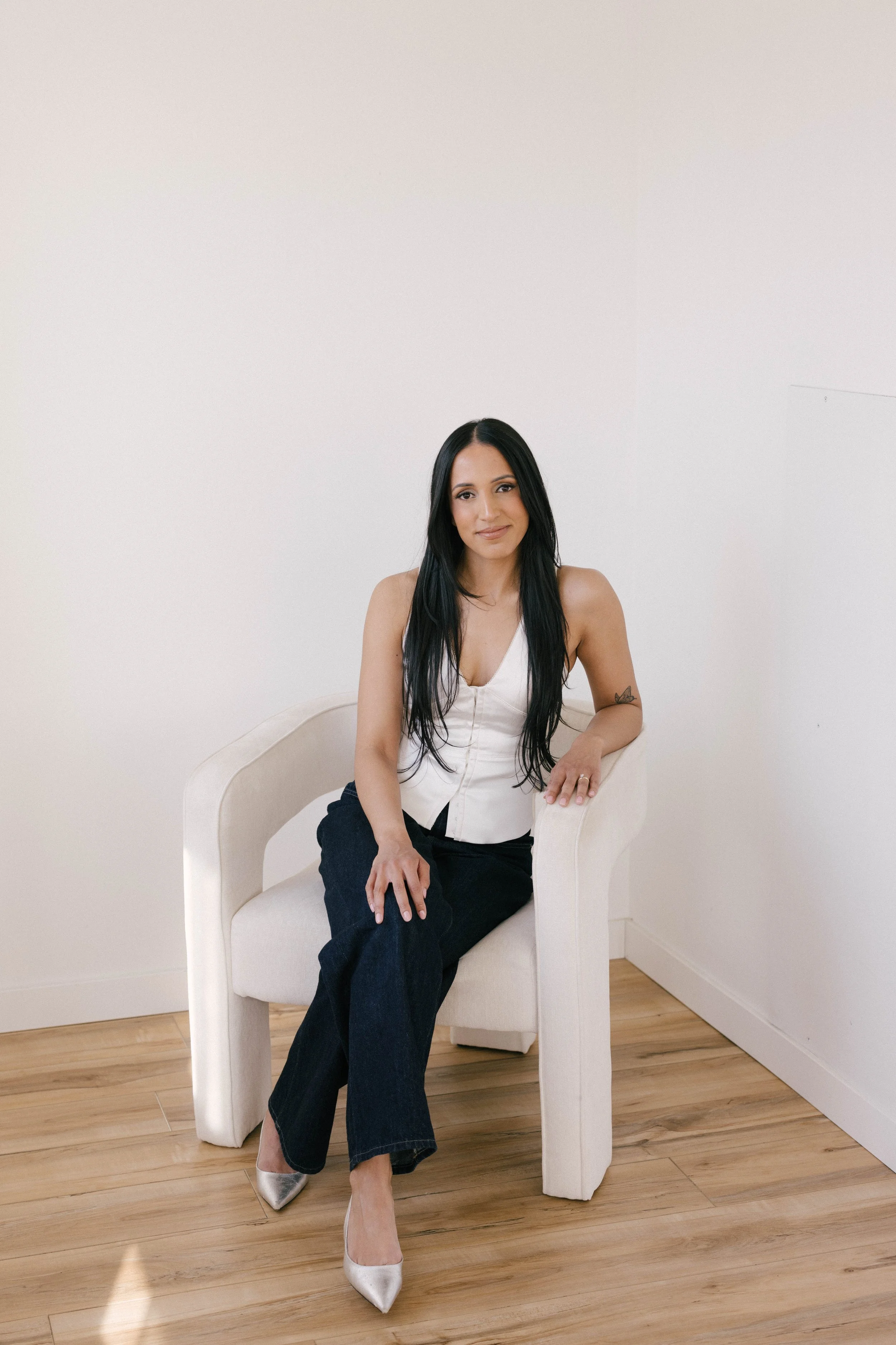 A woman with long dark hair wearing a black tshirt and black pants, sitting white and steel minimalistic room with hardwood floors and a white wall, near a window with sheer white curtains.
