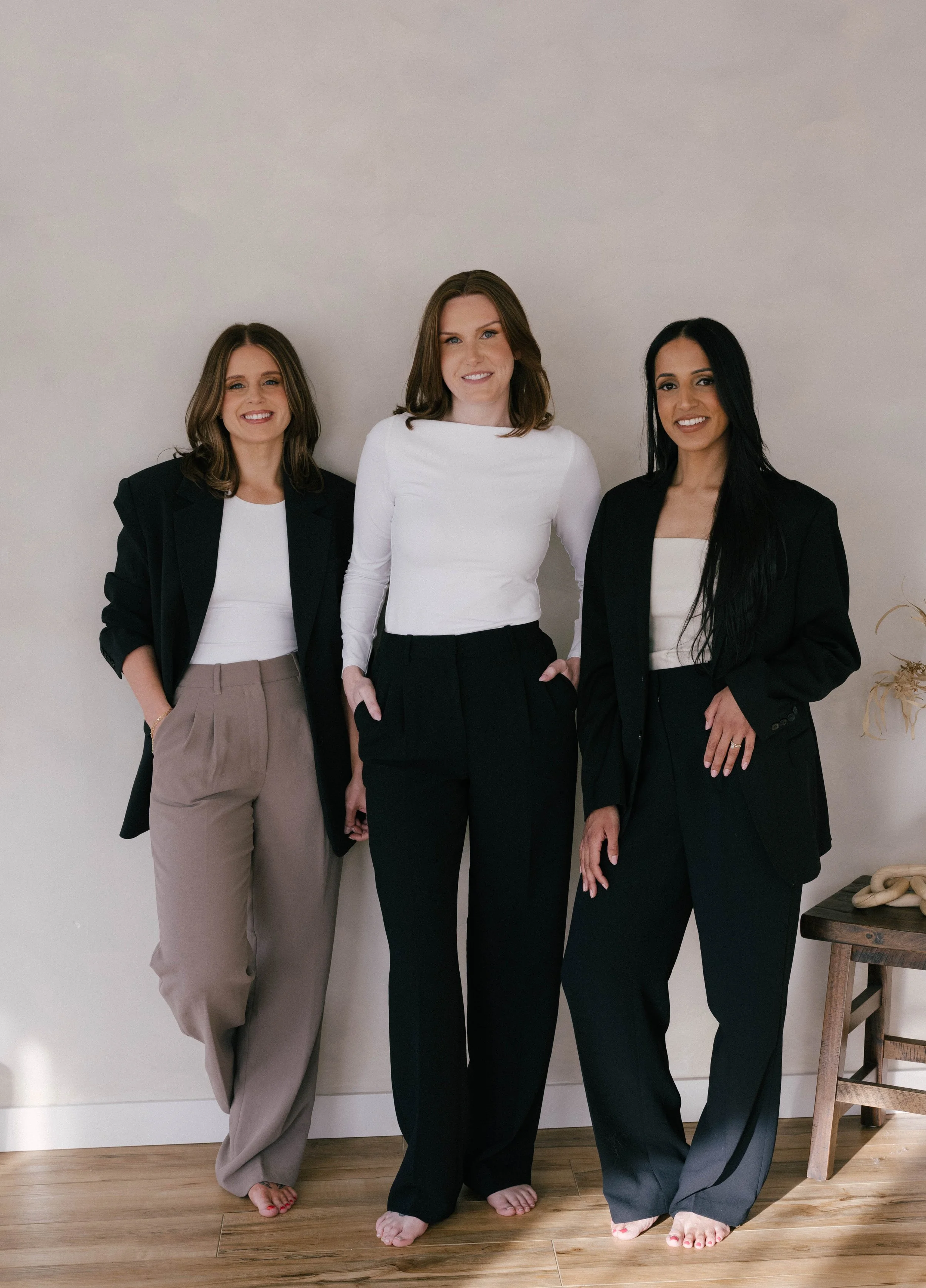 Three women smiling at the camera, leaning against a neutral coloured wall, dressed in black and white business attire