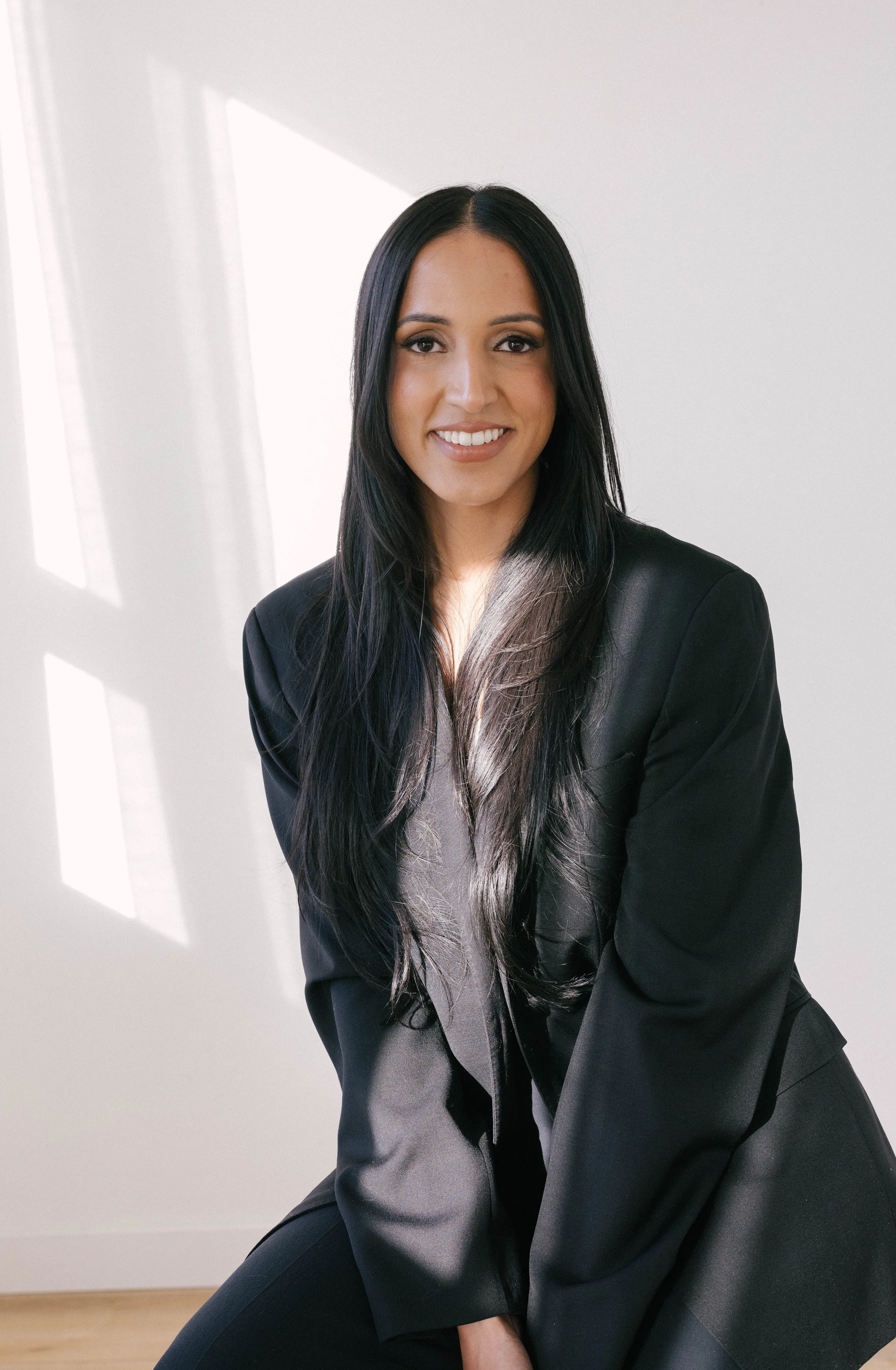 A woman with long black hair and a black blazer smiling, sitting in a well-lit room.