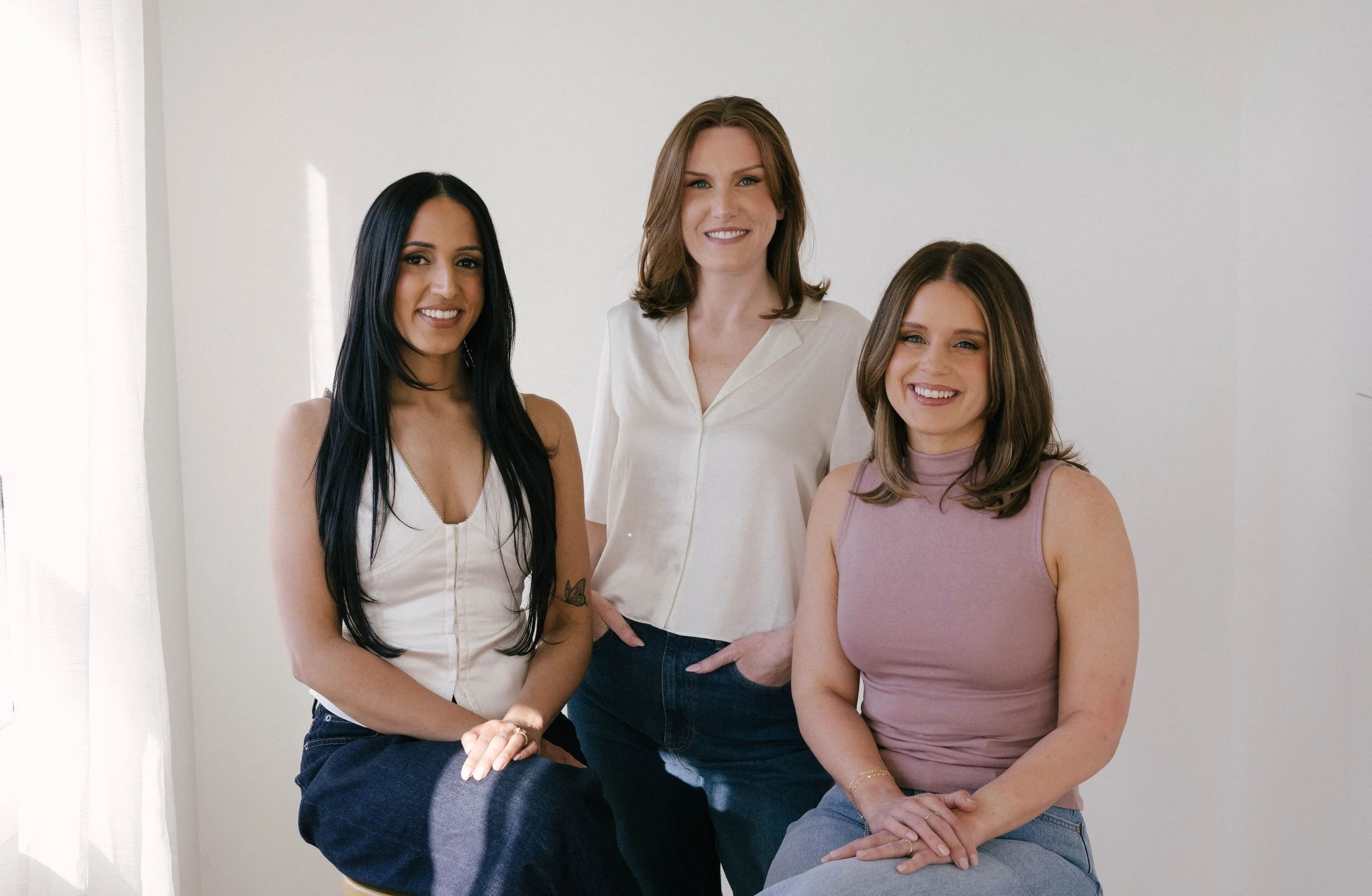 Three women smiling, two sitting on stools and one standing behind with hands in pocket. A white wall is seen in the background with some light shining through.