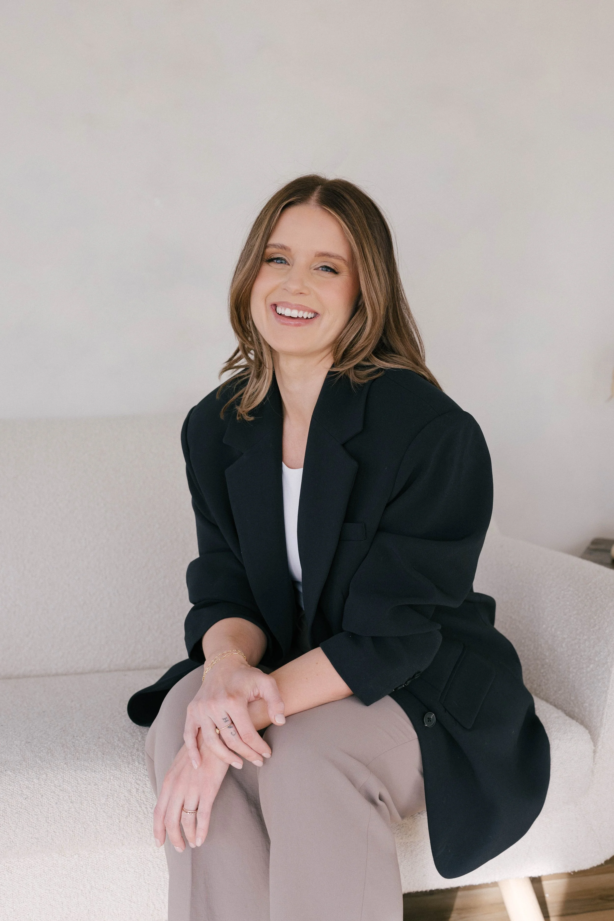 A woman with shoulder-length brown hair, wearing a black blazer and beige pants, smiling and sitting on a cream-colored sofa in a minimalist room.