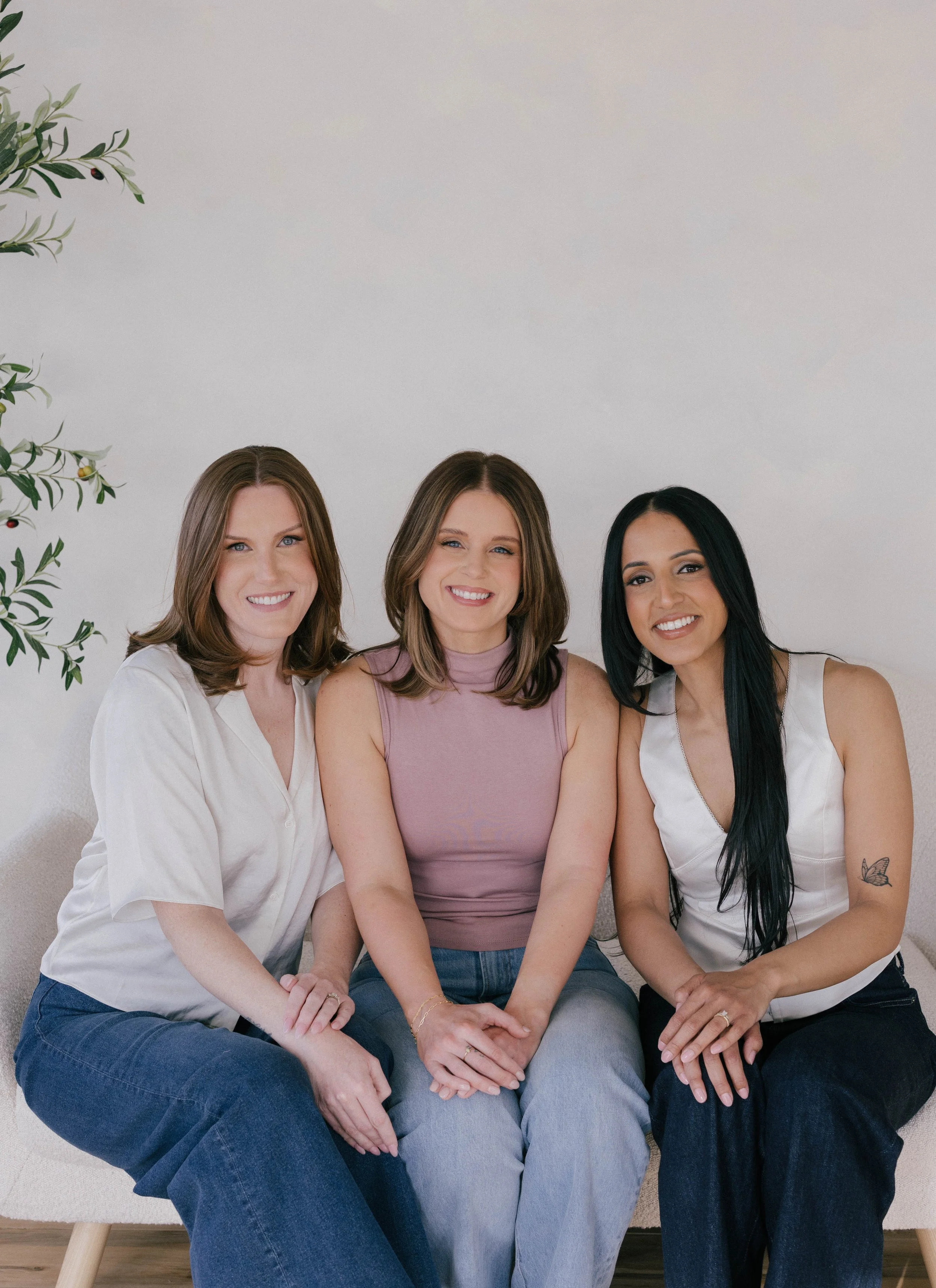 Three women posing together on a white couch in a brightly lit room, smiling at the camera