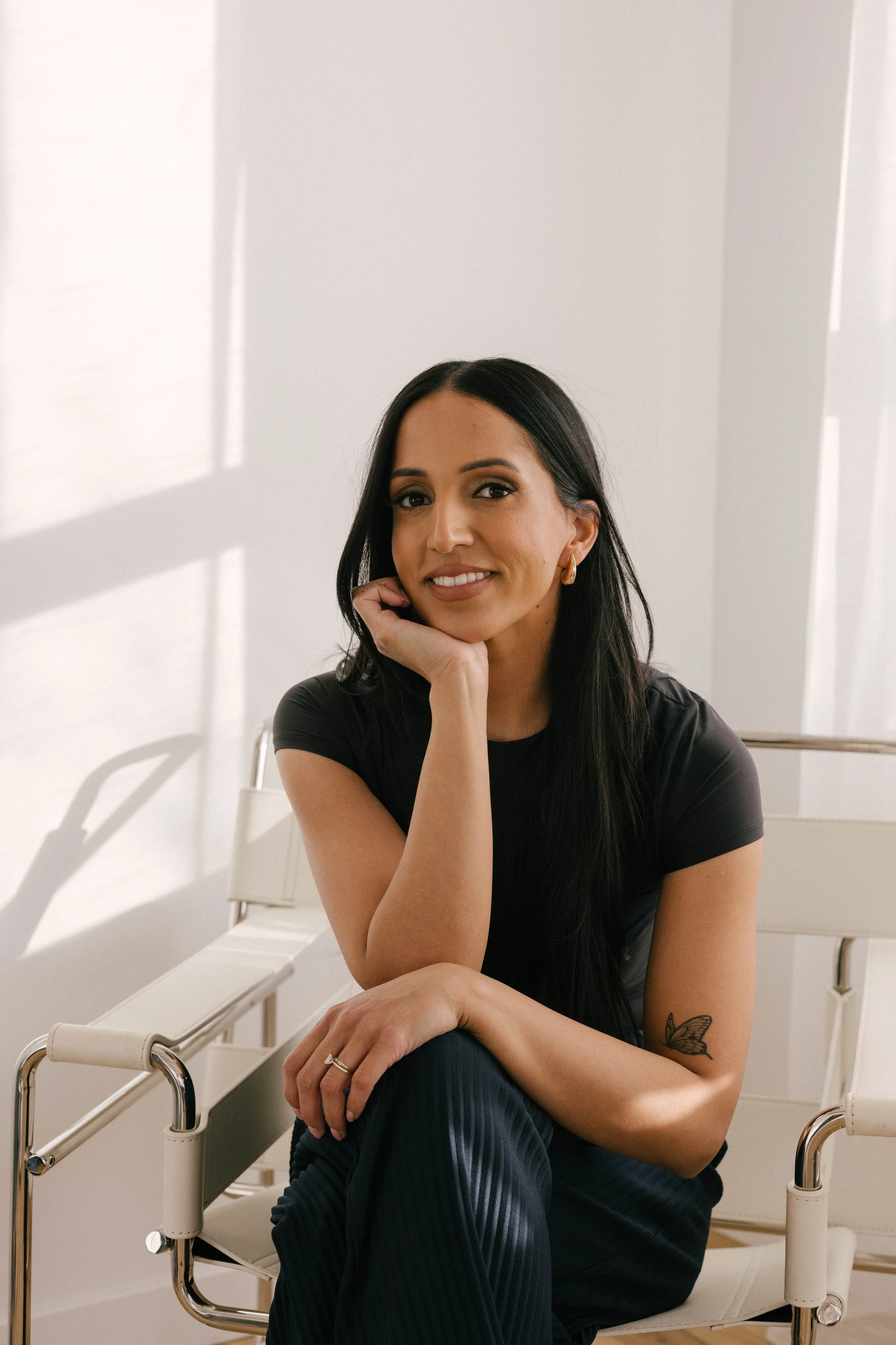 A woman with long dark hair wearing a black tshirt and black pants, sitting white and steel minimalistic room with hardwood floors and a white wall, near a window with sheer white curtains.