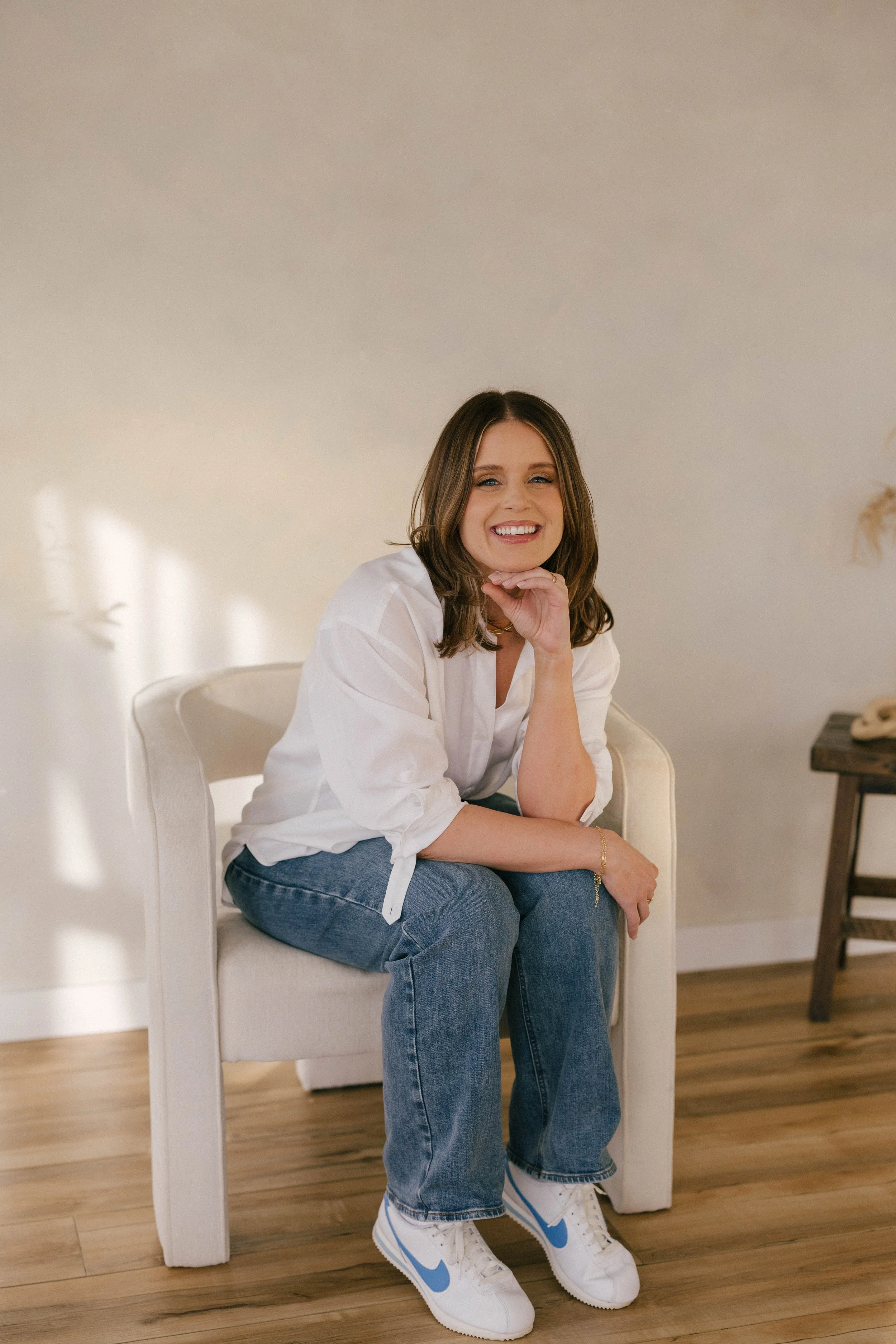 A woman sitting on a white chair, wearing a white blouse, blue jeans, and white sneakers with a blue Nike swoosh, smiling, with her hand resting on her chin, in a bright room with wooden flooring.