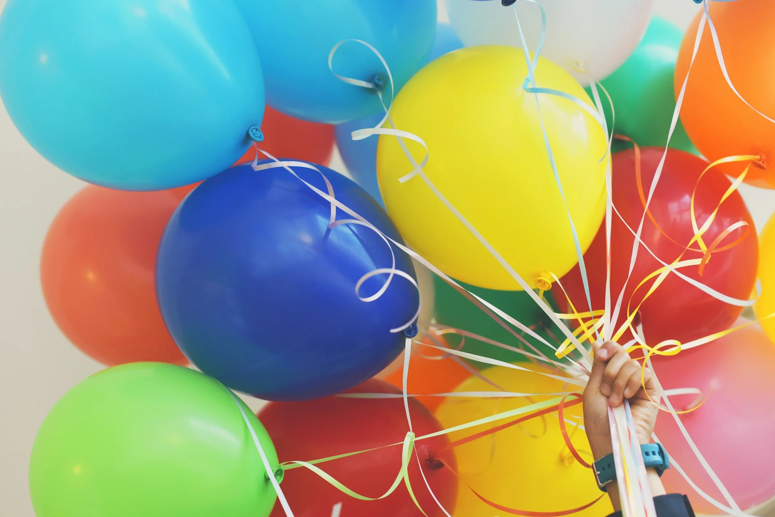A bunch of colorful balloons including blue, green, yellow, and red, held by a hand with wristbands, against a light background.