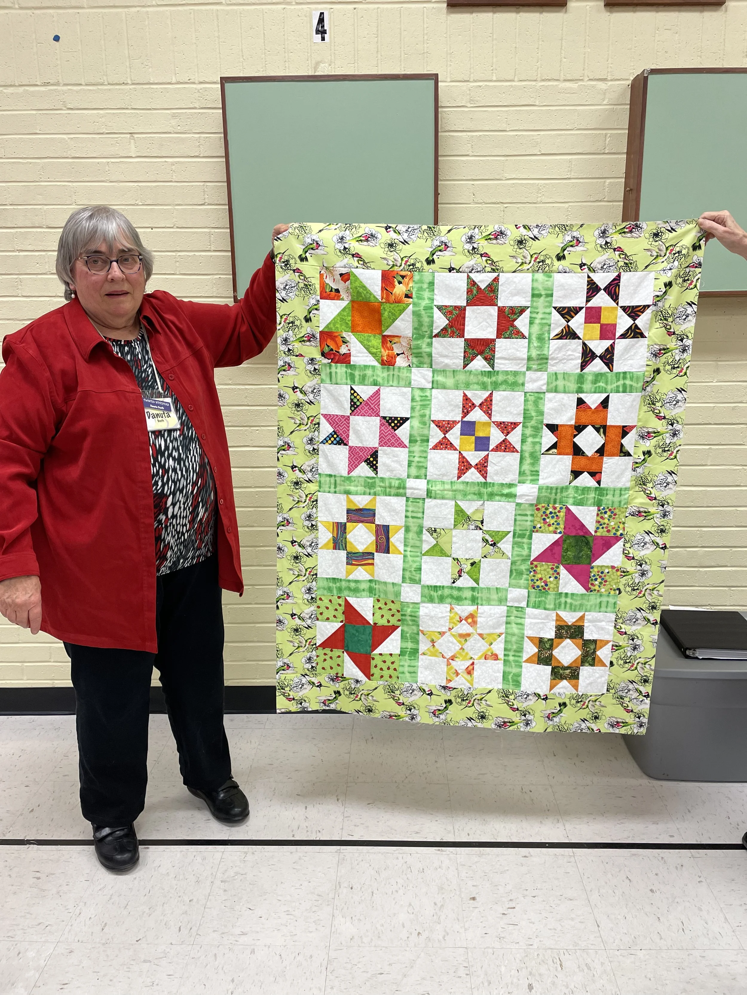 Person holding a colorful patchwork quilt with various geometric patterns in front of a classroom wall.
