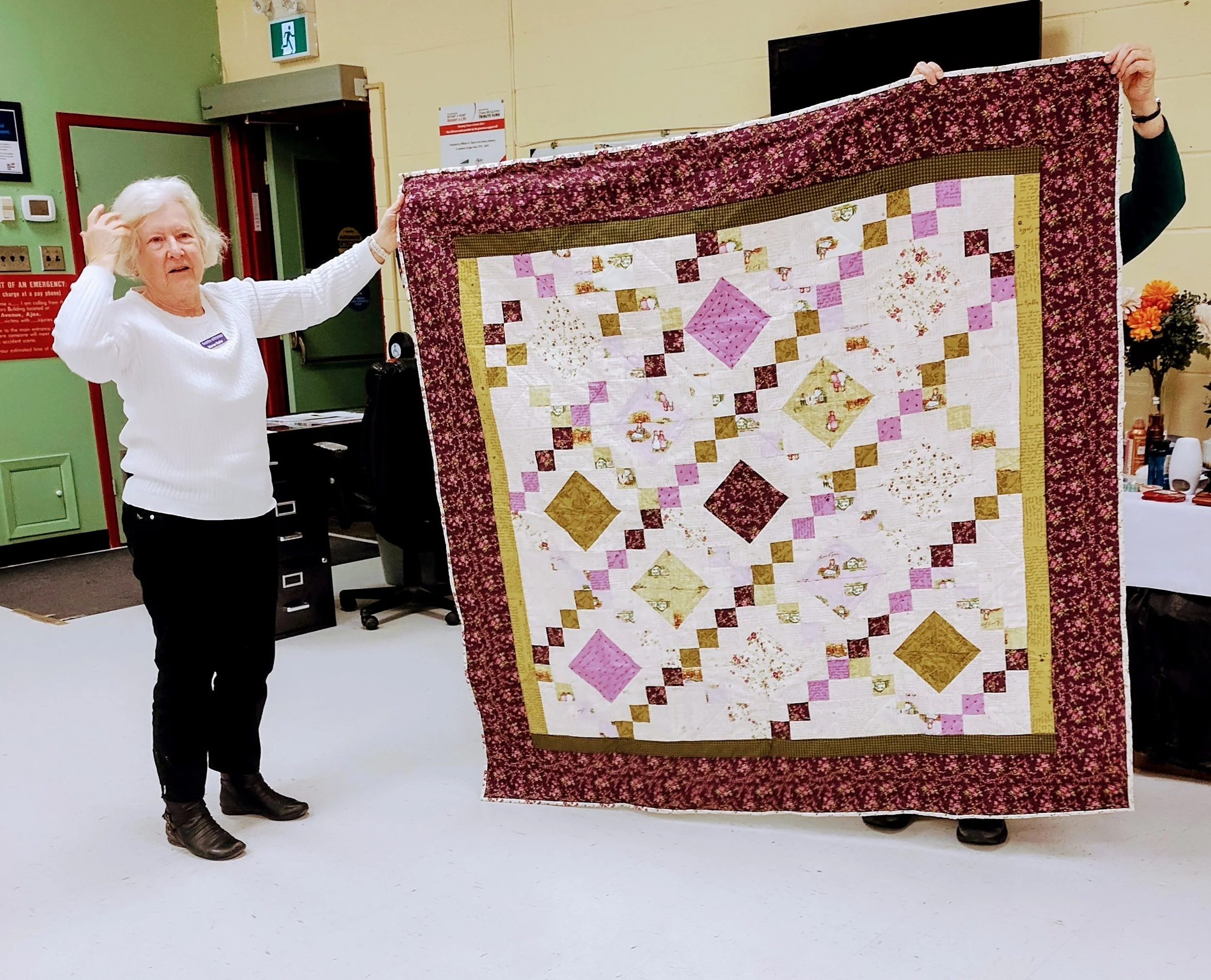 A woman holding a colorful patchwork quilt with floral and geometric patterns in a room with tables and chairs.